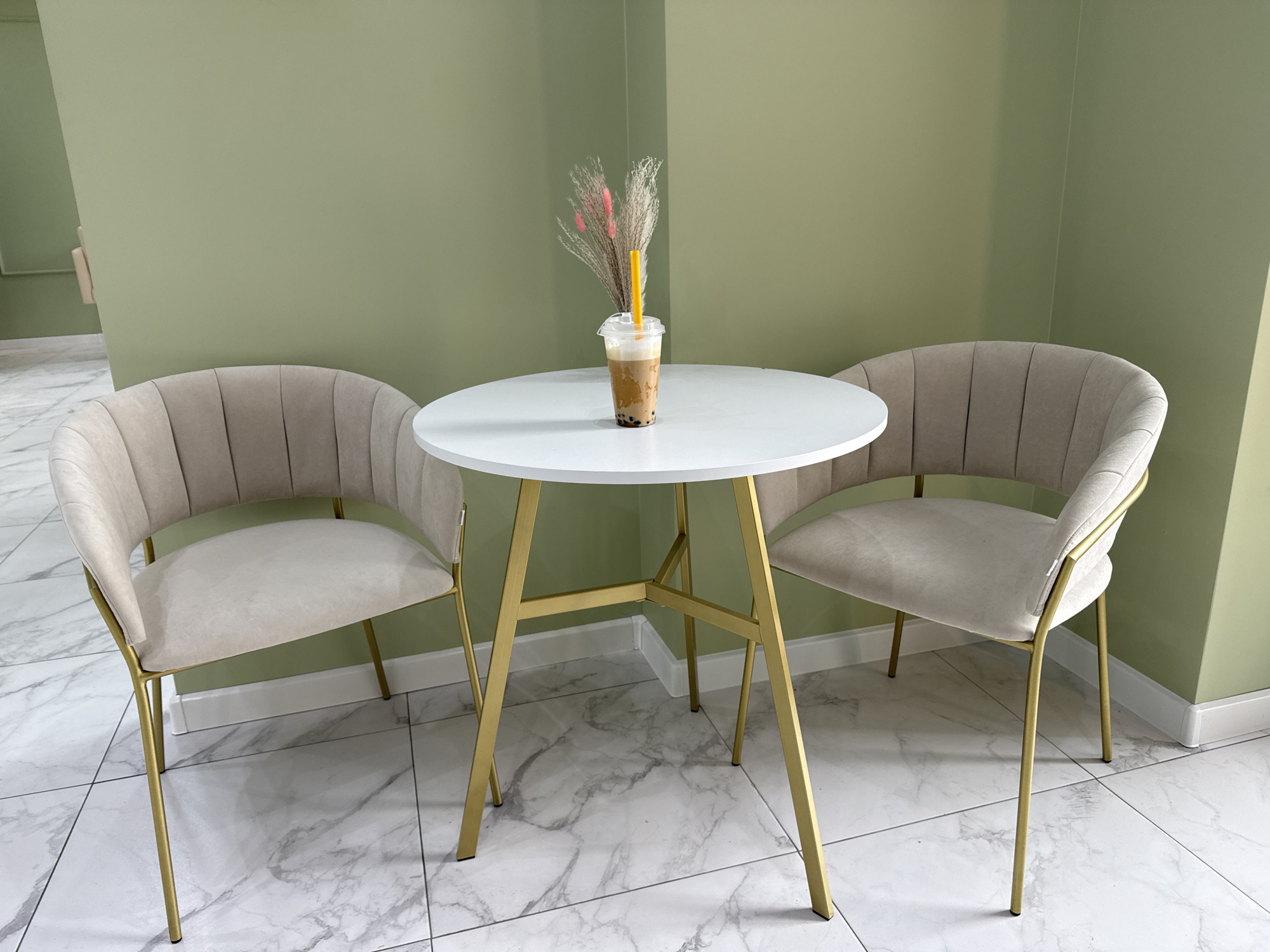 The image depicts a modern cafe setting in Moscow, Russia. The scene features a round, white table with a gold metal frame, centrally positioned between two elegant, upholstered chairs. The chairs are a neutral beige color with plush, channeled seating and delicate gold metal legs that complement the table's base.
On the table, a clear plastic cup holds a beverage, likely a bubble tea, with dark tapioca pearls at the bottom and a creamy, light brown liquid. A bright yellow straw extends from the cup, topped with a small arrangement of dried decorative grasses, some with subtle pink accents.
The background is a soft, muted green wall, creating a serene and contemporary atmosphere. The floor is tiled with a white marble pattern, adding to the sophisticated aesthetic. The lighting suggests it is daytime, with soft, diffused light illuminating the space. There are no people visible in the image, and no text is present. The overall impression is one of a calm, stylish, and inviting corner of a cafe.