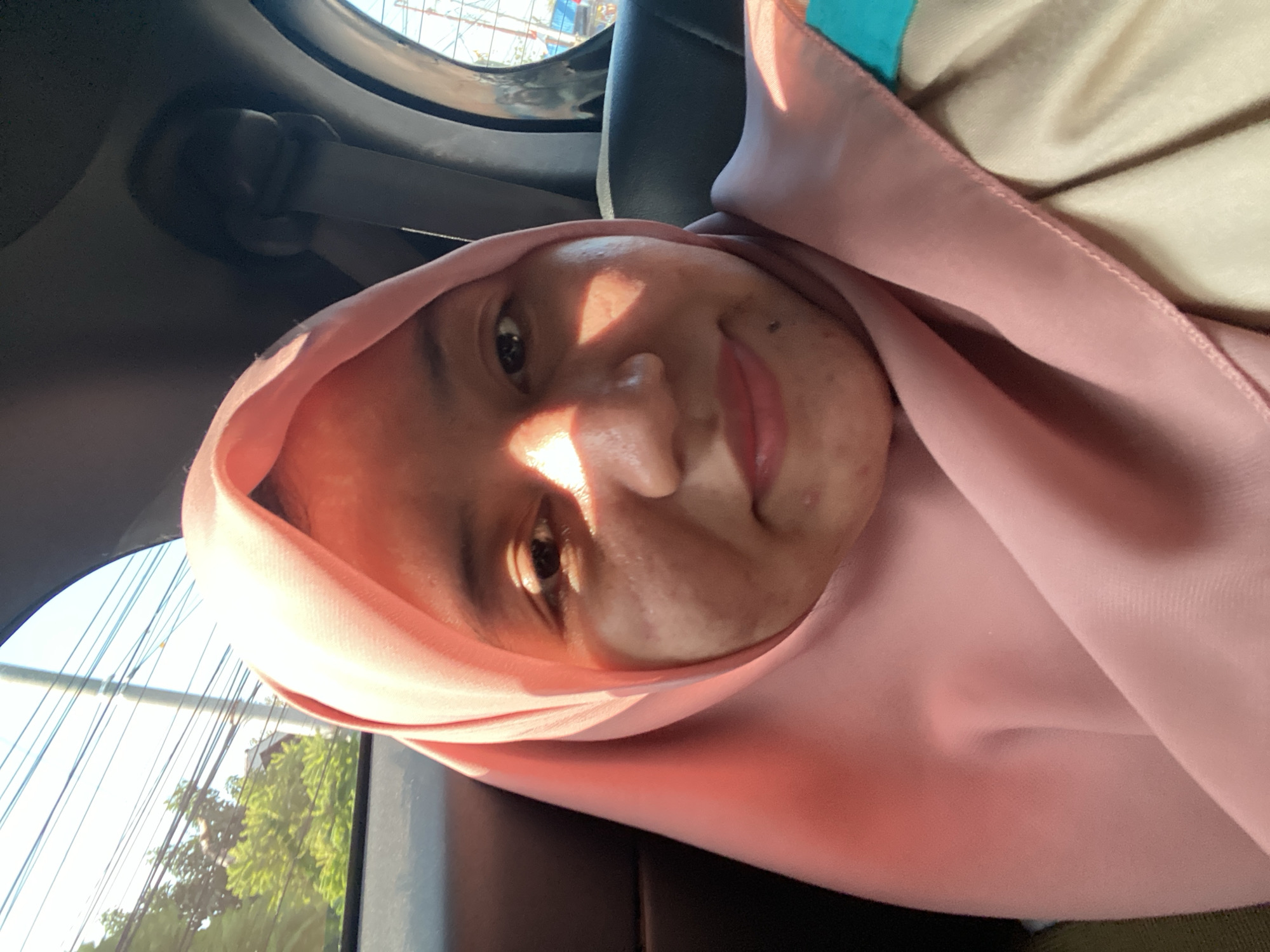 The image is a selfie taken from inside a car, likely in Astambul Kota, Indonesia. A young woman wearing a pink hijab is visible. Sunlight illuminates her face, creating strong highlights and shadows. She is looking directly at the camera with a slight smile. Outside the car window, trees and electrical wires suggest an urban or suburban environment. The interior of the car, including a seatbelt, is also visible. The overall impression is of a casual, candid moment captured during daylight.