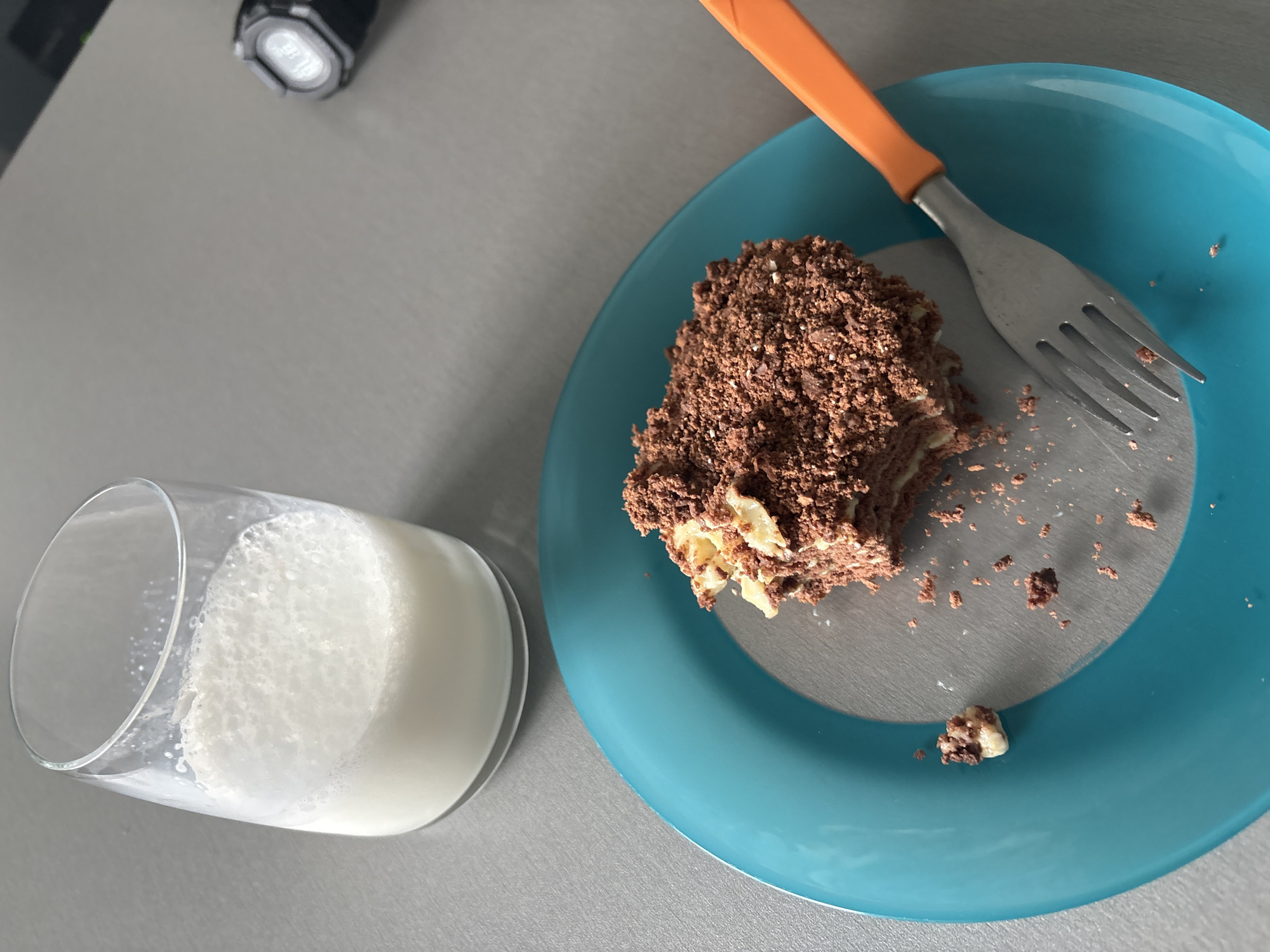 A top-down, eye-level shot reveals a partially eaten chocolate cake on a blue plate, accompanied by a glass of milk and a fork with an orange handle. The cake is situated on the right side of the plate, having had a portion removed. The fork rests on the plate, its tines facing towards the cake. The glass of milk is positioned to the lower left of the plate, its contents appearing frothy. A small, black flashlight sits in the upper left corner of the image. The background consists of a plain, light gray surface. The location of the image is unknown, and therefore cannot be identified in Sofia, Bulgaria. There are no people in the image, and no interactions are visible. The environment is simple and domestic.