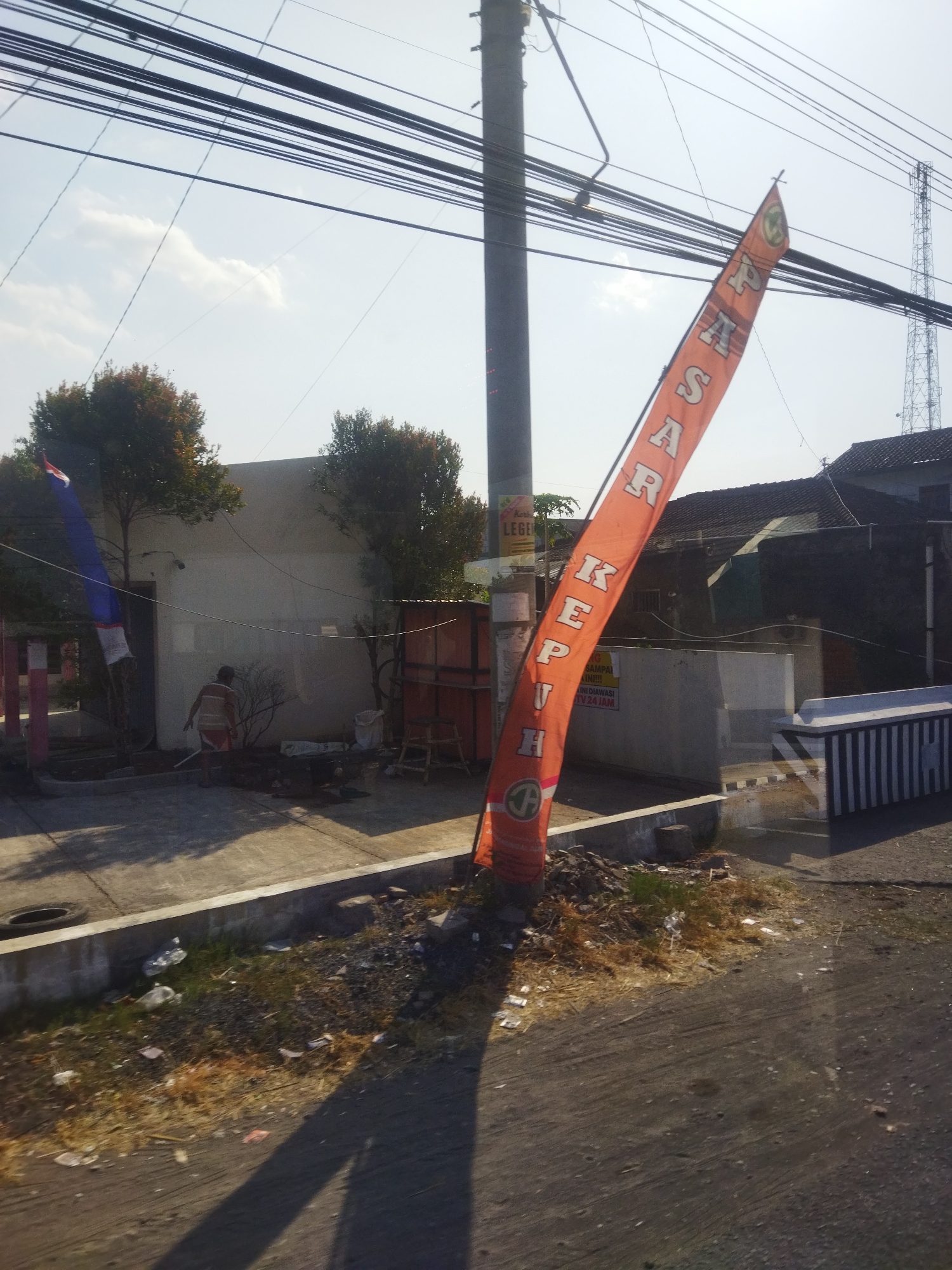 The image captures an outdoor scene in the city of Begajah, Indonesia. In the foreground, an orange banner with white lettering is prominently displayed, angled towards the right side of the frame. The banner appears to have text written vertically in Indonesian, with some letters partially obscured. To the left of the banner is a tall, grey utility pole with numerous black electrical wires running horizontally across the upper portion of the image. Further left, a flag with red and blue stripes is visible, attached to a white pole.

Behind the banner and utility pole, a building with a light-colored facade stands. Two trees with reddish-brown foliage are situated in front of this building. A person wearing a striped shirt and shorts is walking away from the viewer towards the building.

To the right of the banner, a residential structure with a dark tiled roof is visible. In front of this structure, a section of a grey and white striped barrier runs horizontally. The ground in the foreground is paved, with a concrete curb and a patch of dry grass and debris on the left. The overall environment suggests a somewhat neglected urban or semi-urban area, with scattered litter and dry vegetation. The lighting indicates it is daytime, with shadows cast on the ground, suggesting the sun is positioned to the left.