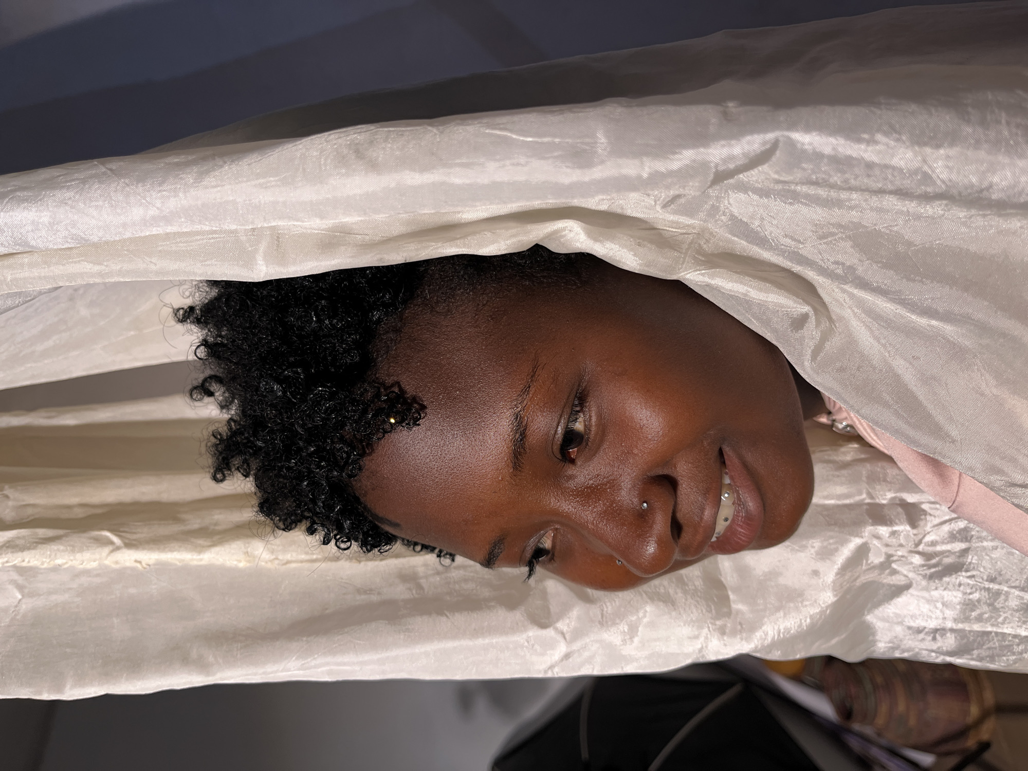 A close-up shot shows a young Black woman's face from the cheekbone upwards, framed by flowing, sheer white fabric. Her curly, dark hair is visible around her forehead and temples. She is looking off to her left with a gentle smile, revealing teeth adorned with small, clear studs. A small, clear stud is also visible on her nose. The fabric appears to be part of a garment or draped material, creating shadows and highlights on her skin. The background is a gradient of soft lighting, transitioning from darker tones on the left to a muted blue on the right. The scene suggests an indoor setting, possibly a studio or a creatively lit room. The location in Barakin Akawo, Nigeria, cannot be determined from the visual information alone.