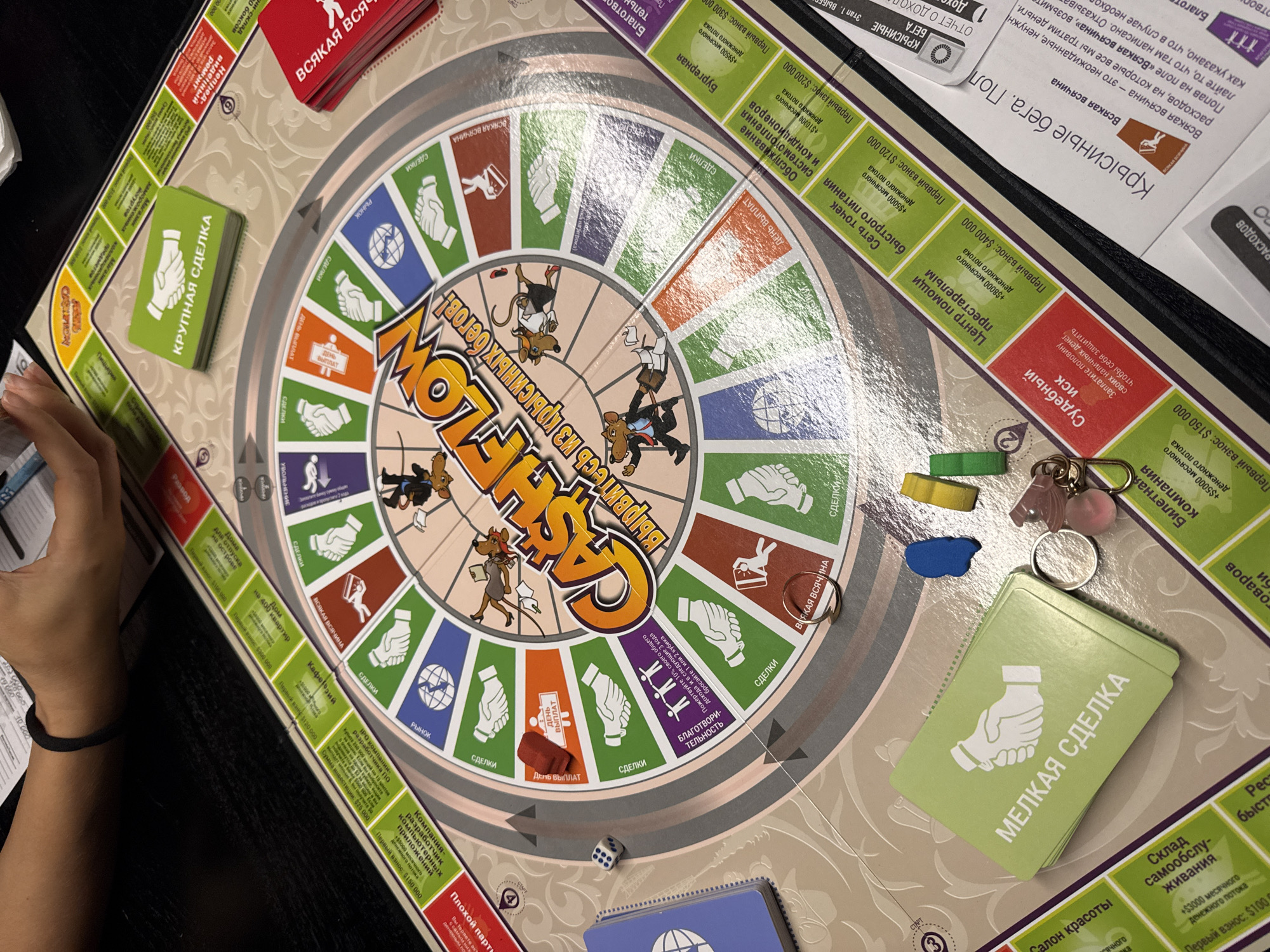 The image shows a Cashflow board game laid out on a dark surface, partially obscured by a person's arm and hand in the lower left corner, with their bracelet visible. The game board is round with a colorful center depicting rats in business attire running in a circle, while the outer ring features various deal opportunities and expenses represented by icons and text. There are stacks of cards labeled "КРУПНАЯ СДЕЛКА" (large deal) in green, "МЕЛКАЯ СДЕЛКА" (small deal) in light green, and "ВСЯКАЯ ВСЯЧИНА" (all sorts of things) in red. Game pieces, including colored markers, a ring, and a set of keys, are scattered near the cards. A single die shows a "1". Parts of the surrounding game materials, including text descriptions and a financial report, are visible around the board. The location is not depicted in this image.