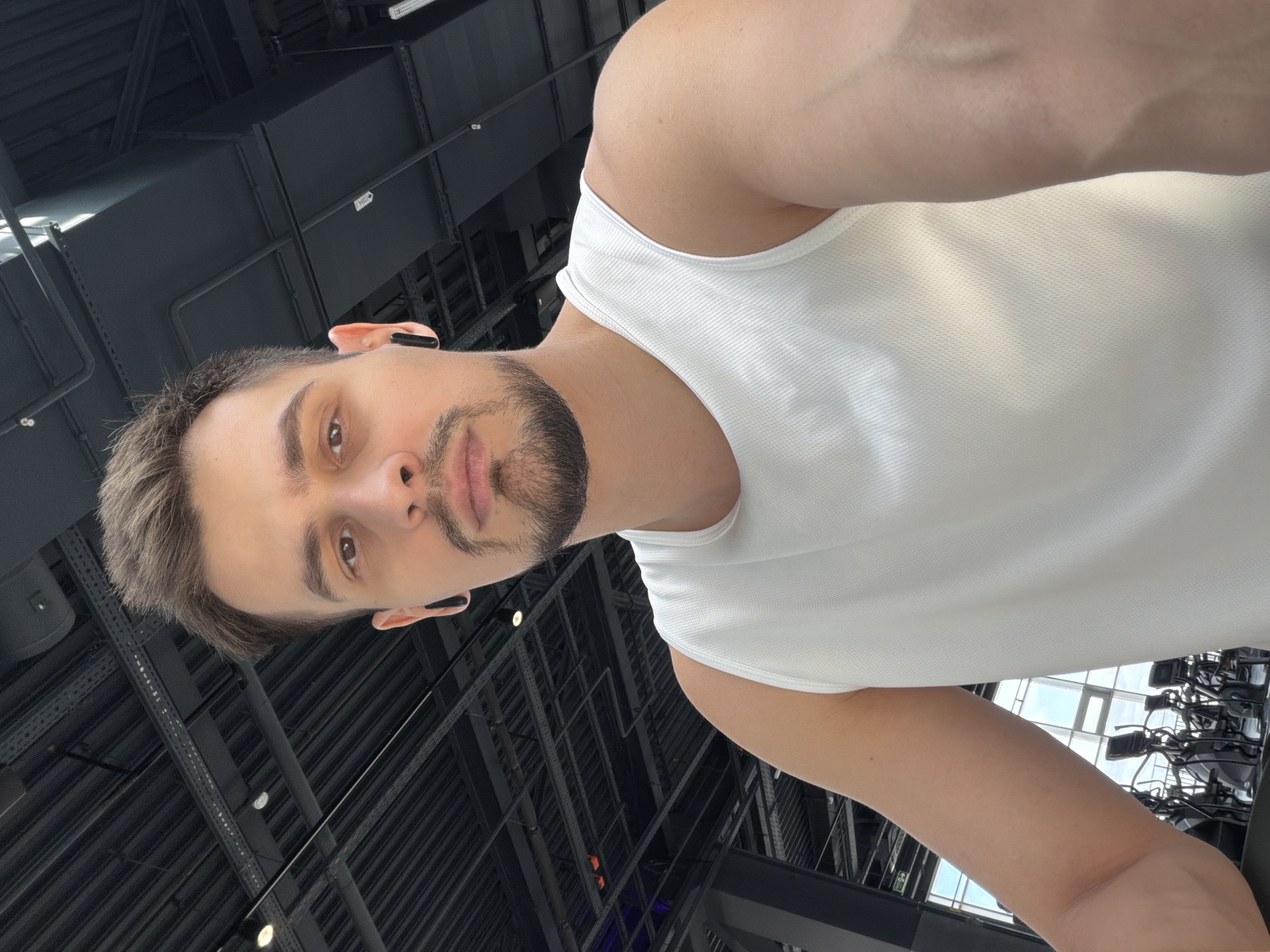 A man is wearing a white tank top. He has short, dark hair and a neatly trimmed beard. He has earbuds in his ears. The ceiling above him has visible metal frameworks and light fixtures. There are workout machines behind him.
The image does not give enough information to identify a specific location in Krasnoyarsk, Russia.