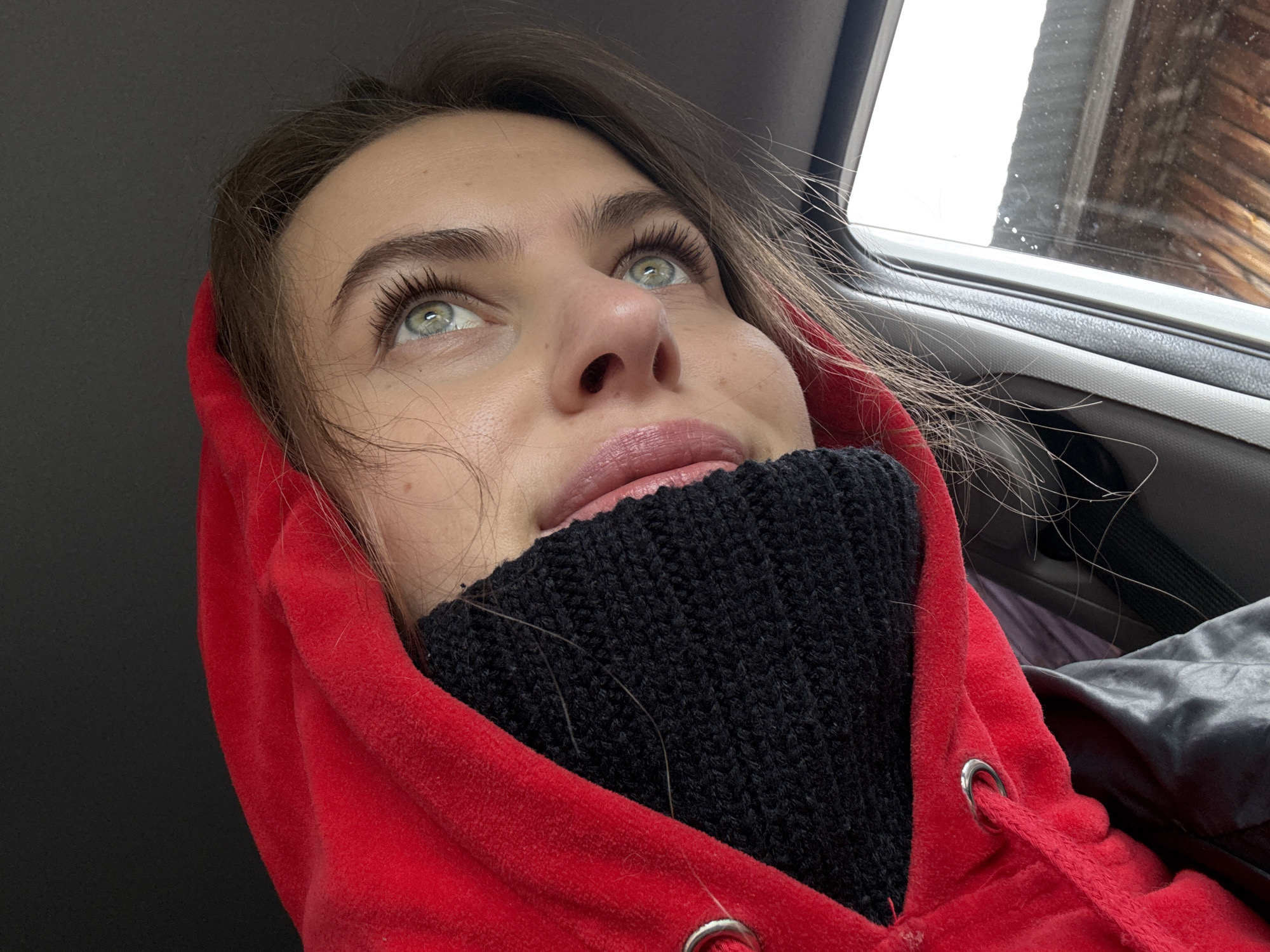 A close-up, eye-level shot captures a young woman seated inside a vehicle, likely a car, looking upwards and to her right with a contemplative expression. Her fair skin, striking light green or hazel eyes with prominent eyelashes, and well-defined eyebrows are central to the composition. Her lips are naturally pink and slightly parted.
She is dressed in a bright red hooded sweatshirt, possibly made of velour or fleece, with a silver metal eyelet visible near the bottom right. Underneath, a black knitted turtleneck or snood covers her chin and neck, suggesting a need for warmth. Her medium brown hair is slightly dishevelled around her face and shoulders, adding to a candid feel.
Through the car window on the right, a bright, possibly overcast or snowy sky is visible above a rustic wooden structure, which could be a house or barn. Small white specks on the window might indicate precipitation. The visible car interior includes white and dark grey plastic trim. A grey or black satin-like fabric is also visible in the bottom right corner. The ambient light suggests it is daytime. The scene evokes a moment of quiet observation or reflection, possibly during a journey in the Прикамский region of Russia. There is no visible text.