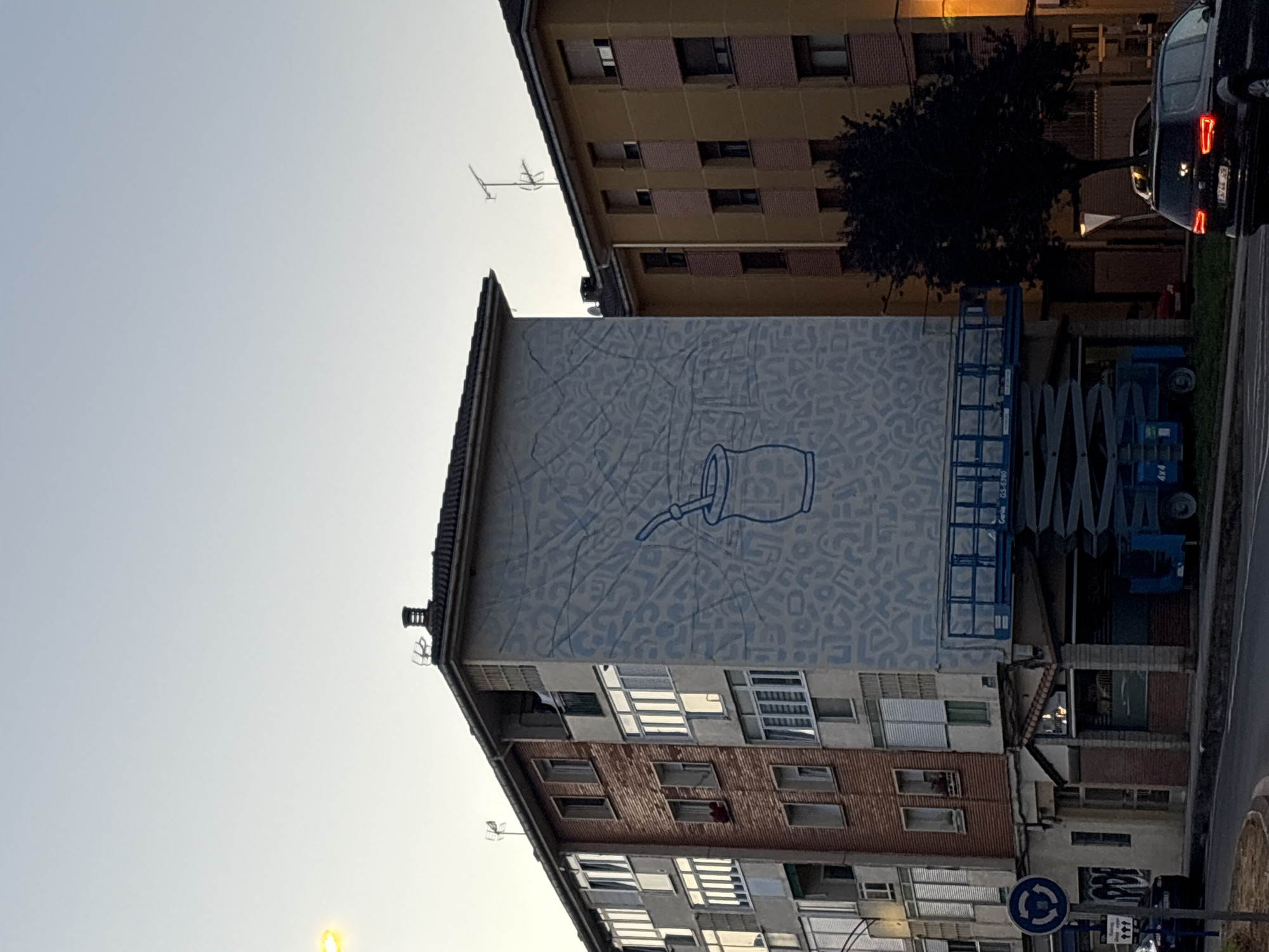 A wide-angle, low-angle exterior shot captures a city scene in Mungia, Spain, at dusk. The sky is a pale, uniform blue, suggesting the end of the day. Dominating the left side of the frame is a multi-story brick and concrete building adorned with a large blue mural. The mural depicts a stylized illustration of a mate gourd with a straw, set against a background of abstract, light blue shapes that resemble lettering or patterns. A blue scissor lift is positioned in front of the mural, suggesting ongoing work or maintenance.
To the right of the muraled building is a smaller, pale yellow building with brown and pink accents. This building also has multiple windows, some with shutters. A dark silhouette of a tree stands in front of the yellow building. The foreground shows a street with a paved surface and a sidewalk. A dark-colored SUV with its taillights illuminated is parked on the right side of the street. A blue and white circular traffic sign with three arrows pointing clockwise is visible on a pole to the left of the muraled building, near a graffiti-covered wall. The overall atmosphere is one of urban residential architecture.