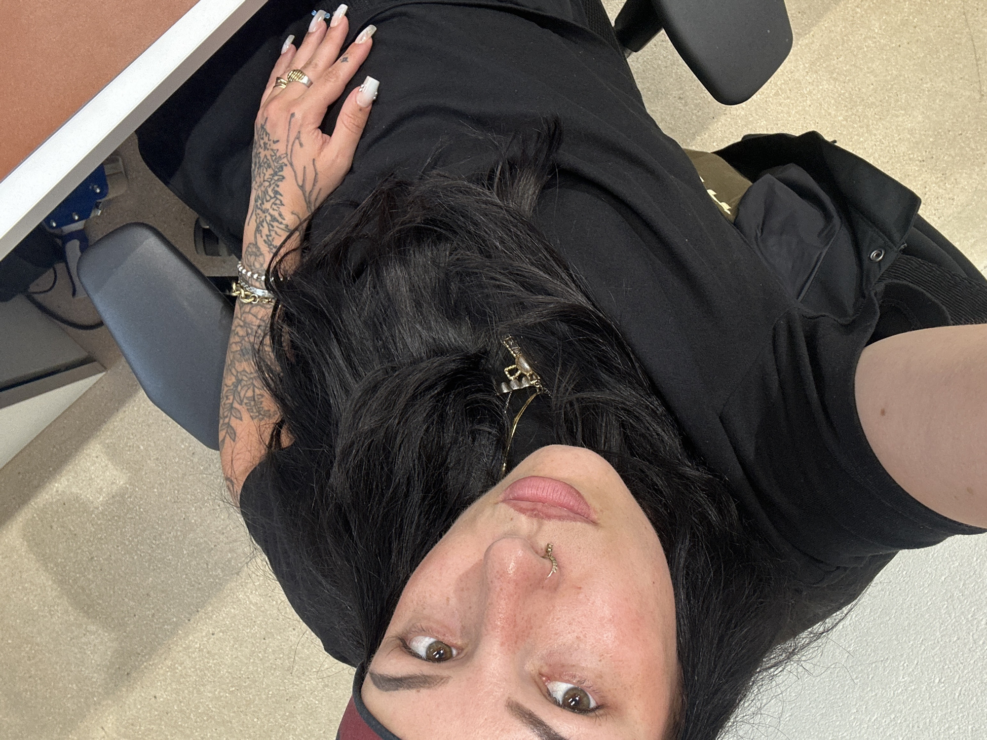 A selfie-style, close-up photograph shows a person with long, dark hair, a nose piercing, and visible tattoos on their left hand and arm. They are wearing a black top and a gold necklace. Their left hand, adorned with rings and bracelets, rests on their lap. The background includes a desk with a white edge and a chair. The floor appears to be light-colored and uniform. No evidence suggests the location is Paris, France.