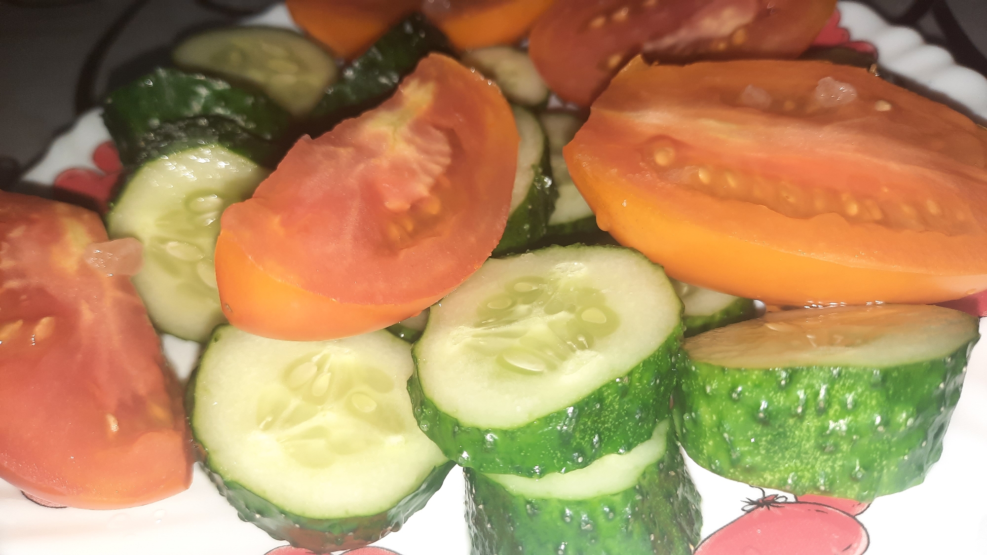 A close-up, overhead shot shows a plate of sliced tomatoes and cucumbers. The tomatoes are red and orange, cut into wedges and halves, revealing their seedy interiors. The cucumbers are sliced into rounds, showcasing their green skin and pale, seedy flesh. Some of the vegetables appear wet, suggesting they might be part of a salad or a fresh preparation. The background is indistinct but hints at a patterned plate with red and white designs. There is no indication of the city of Shklov, Belarus, or any other environmental context.