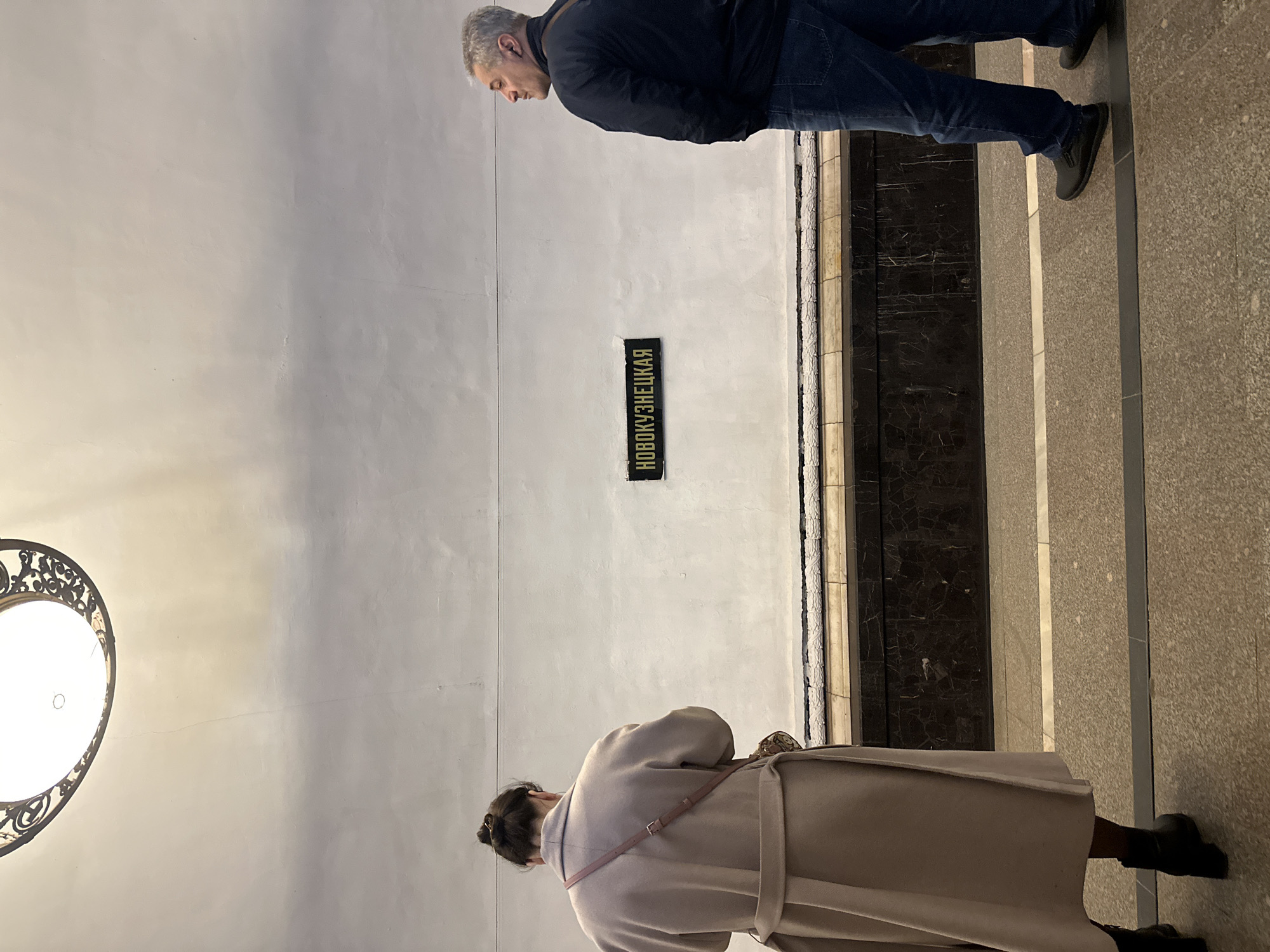 A man and a woman are in the Novokuznetskaya metro station in Moscow, Russia. The man is standing while the woman is looking down. The metro station's name is in Russian on the wall.