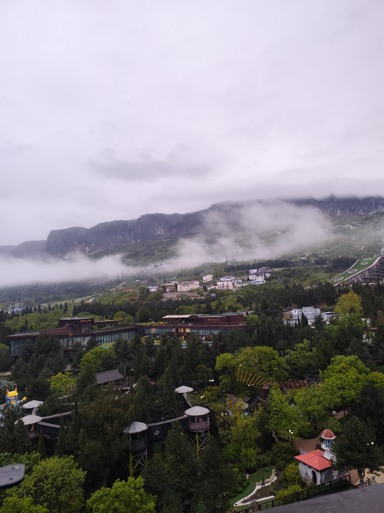 The image shows a view overlooking a mountainous landscape in the city of Симеизский поселковый совет, Ukraine, under a cloudy sky. Mountains are partially obscured by low-lying clouds and fog. In the foreground, a wooded area contains various structures, including lookout towers connected by elevated walkways, and a miniature golf course with a small building featuring a red roof and blue dome. Further back, a modern, multi-story building with horizontal architectural elements and the sign "Mriya Life" is visible amidst the trees. On the mountain slopes behind, several residential houses are scattered. A long, green-roofed building, possibly a hotel or resort, is on the right side of the houses.