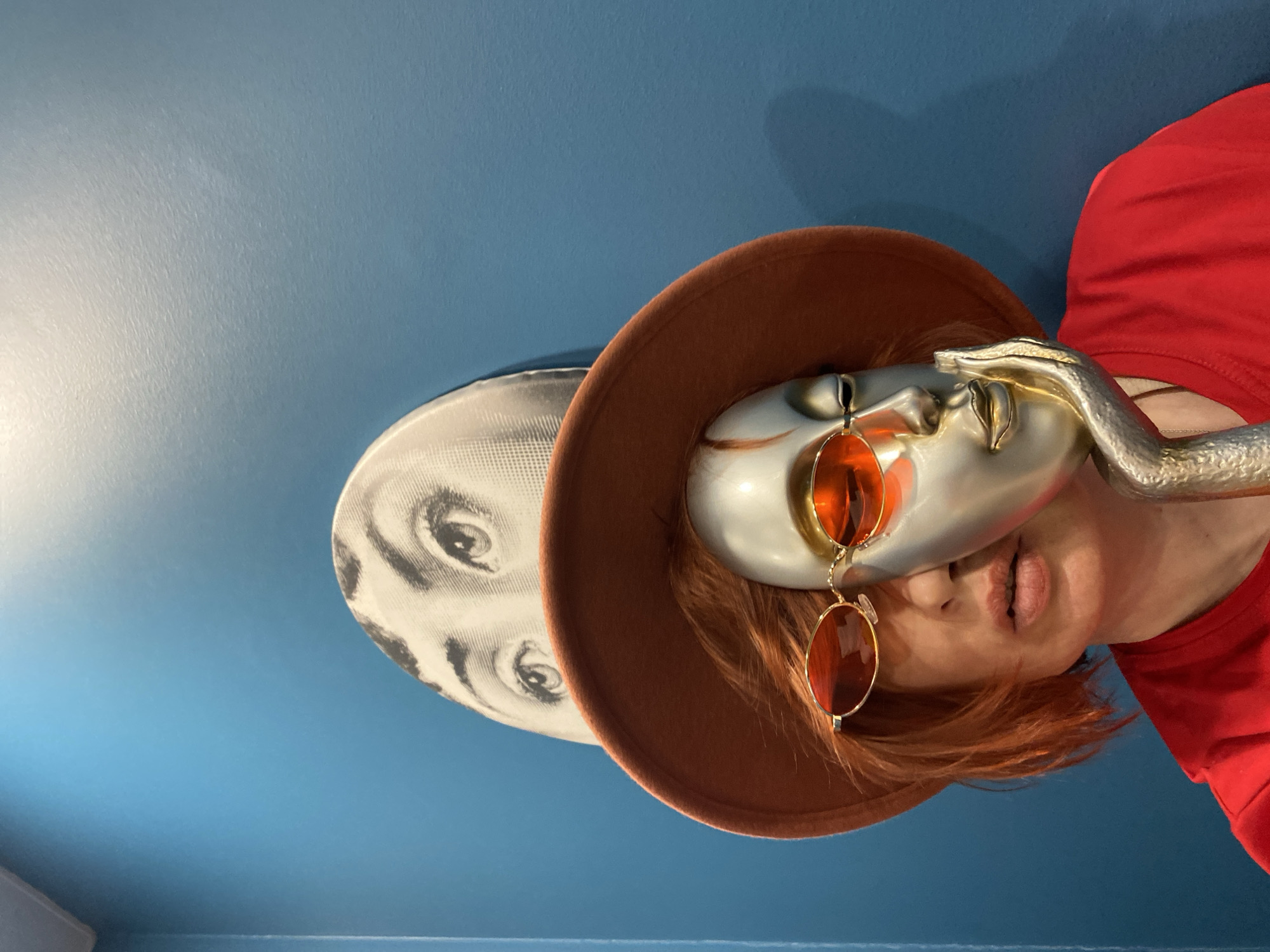 The image shows a person wearing a red shirt and an orange wig. A metal mask covers part of the person's face. The mask is adorned with orange glasses and a brown hat. A silver, arm-shaped sculpture is draped over the person's shoulder. In the background, a white plate with a black-and-white image of eyes is visible. The background is a solid blue. The image does not contain sufficient information to determine if it was taken in Yekaterinburg, Russia.