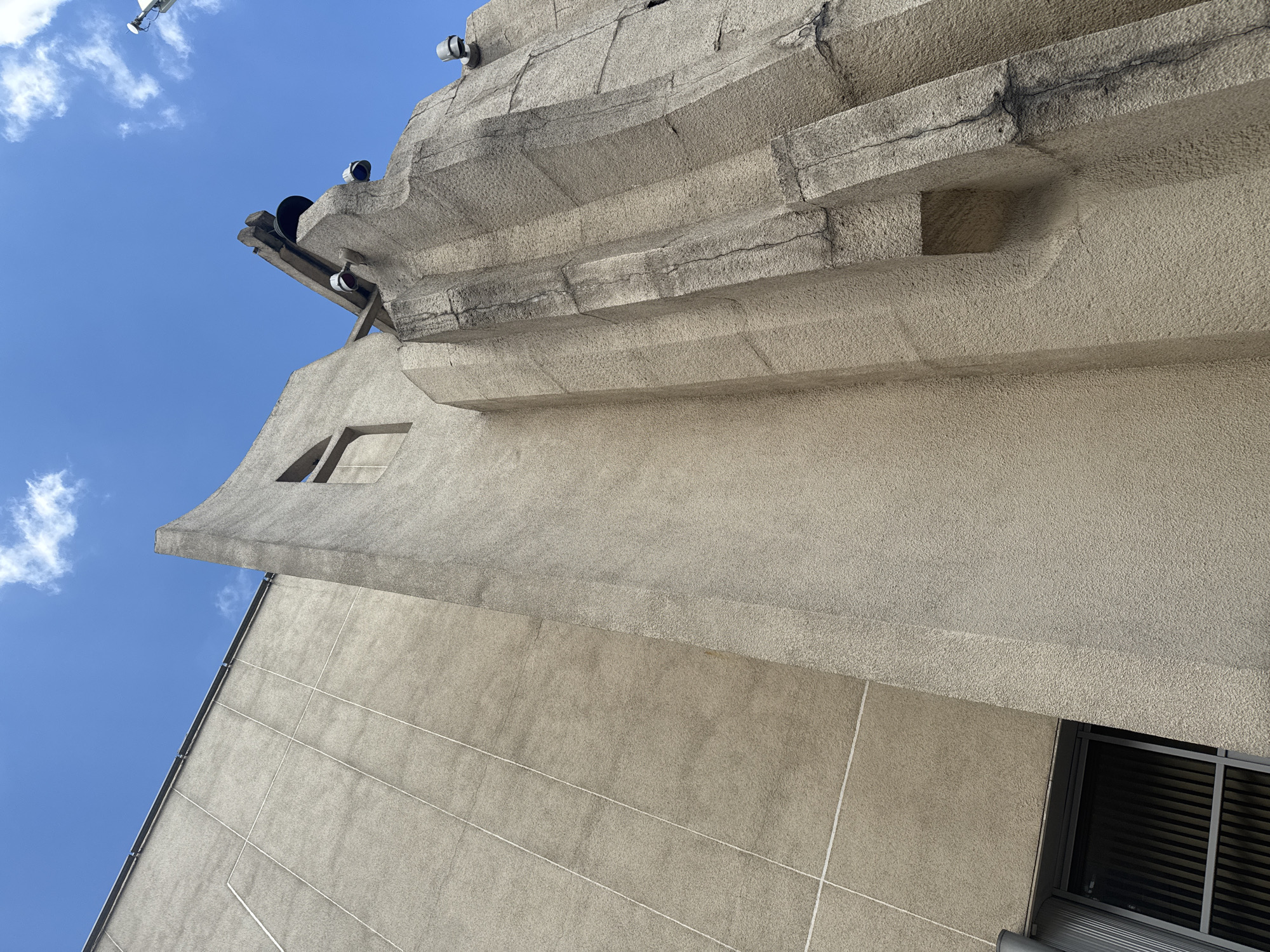 The image shows a low-angle view of a concrete building exterior against a bright blue sky with scattered white clouds. The building has a textured, light beige concrete facade with vertical lines scored into it in some areas, and a more rugged, art-deco-inspired design in other parts. On the right side of the building, there are several protruding architectural elements and what appears to be a bell hanging from a crossbeam, with lights mounted nearby. There is a small, rectangular window visible on the upper portion of the more angular section of the building. Near the bottom left, a dark-framed window with vertical blinds is partially visible. The overall impression is of a somewhat brutalist architectural style. The location is identified as the Moscow Planetarium, located at 5, Sadovaya-Kudrinskaya Street, Moscow, Russia.