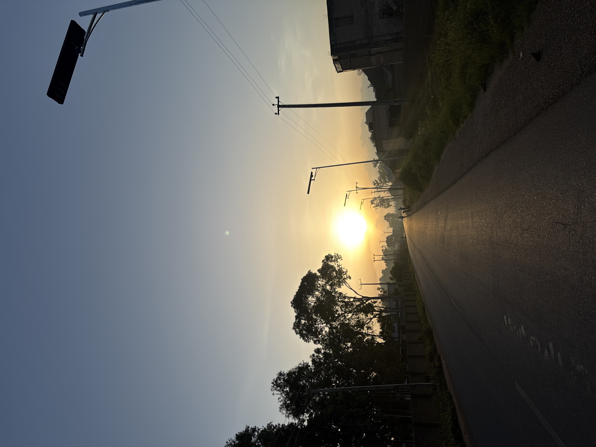 The image depicts a road in Bukuru, Nigeria, at sunrise. The sun, a bright orb, is low on the horizon, casting a warm, golden light across the scene. The road stretches into the distance, with a slight sheen from the morning dew or recent rain. Silhouetted trees line the left side of the road, their branches reaching towards the sky. On the right, there are some buildings and a few streetlights, some of which appear to be modern solar-powered models. A few figures, possibly people, are visible walking on the side of the road in the distance. The sky is a clear, soft blue, transitioning to a pale yellow near the horizon where the sun is located. The overall atmosphere is one of a quiet morning beginning in a semi-urban environment.