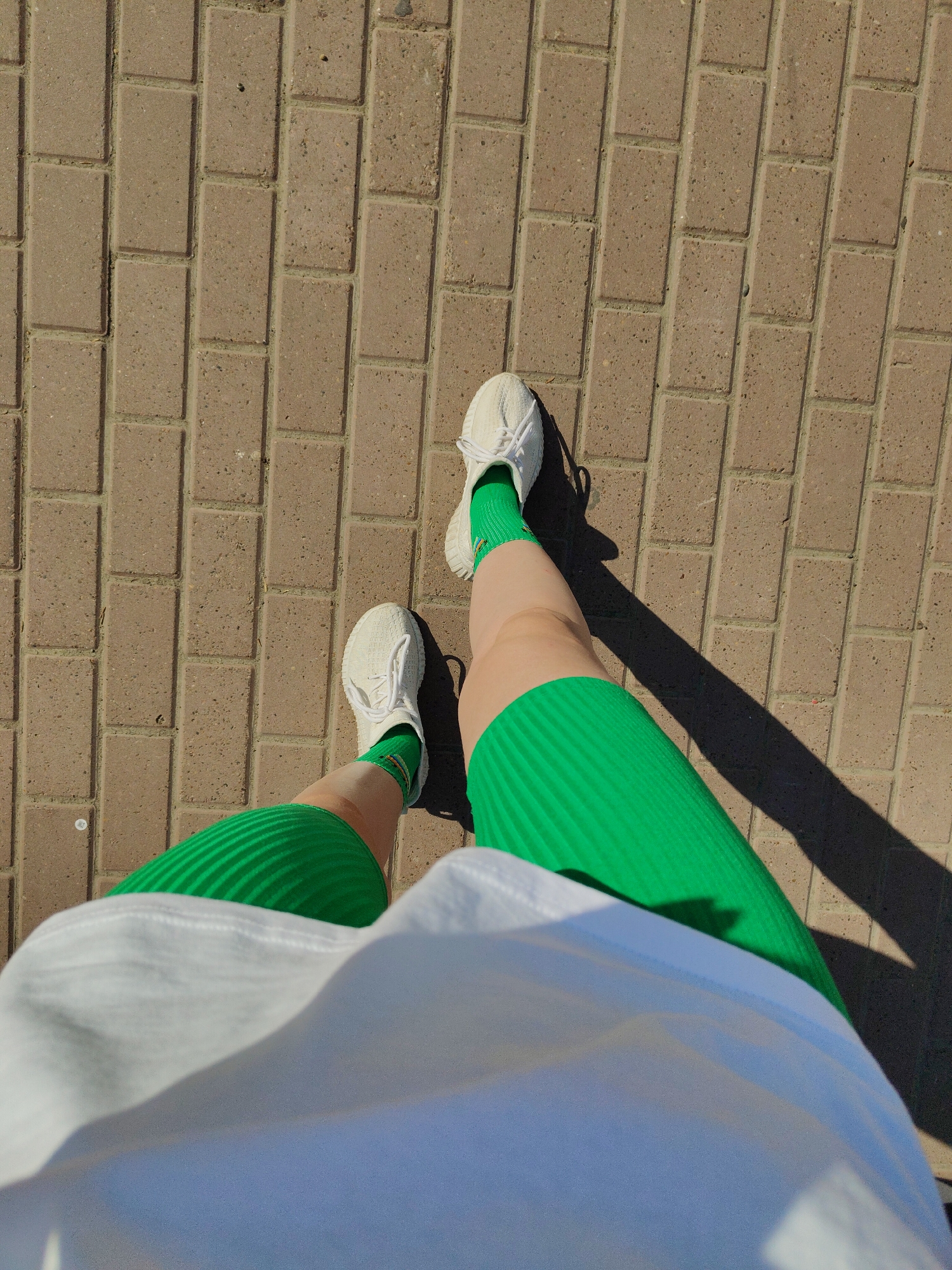 The image shows a person standing on a brick walkway, likely in an urban setting. The person is wearing white sneakers, bright green socks, and a green skirt or shorts, paired with a white top. The perspective is from above, looking down at the person's legs and feet. Sunlight casts shadows on the brick walkway, indicating a sunny day. The bricks are laid in a rectangular pattern.