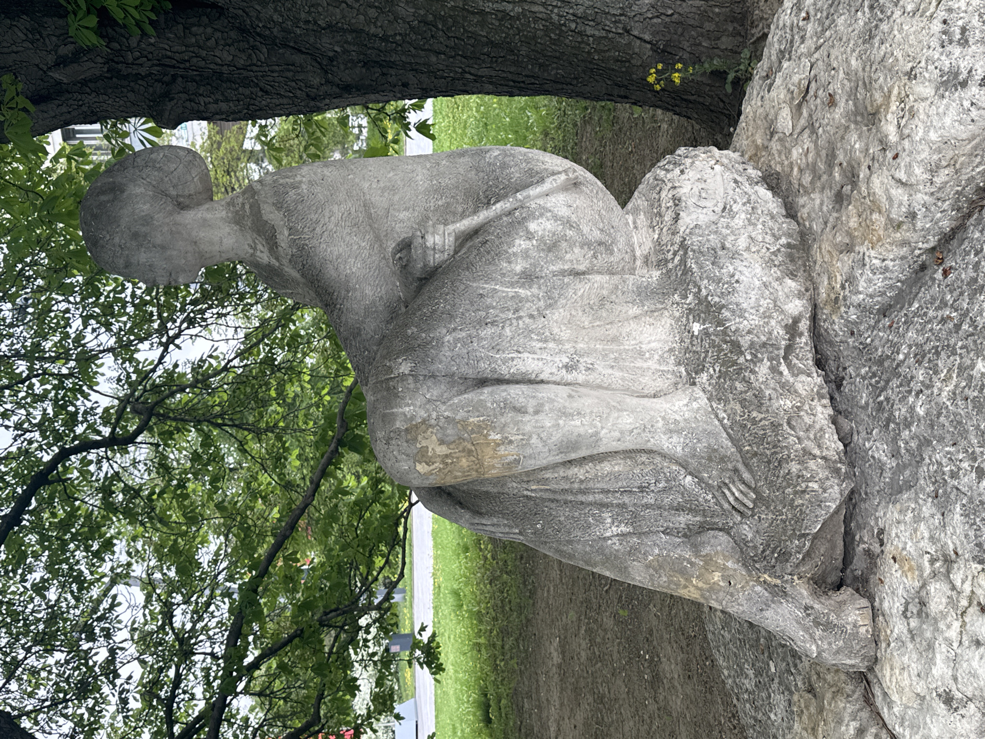The image shows a weathered stone sculpture of a seated, possibly kneeling, human figure. The figure has a simplified, almost abstract form, with a rounded head and indistinct features. The body is draped in cloth. The sculpture is positioned outdoors, nestled between the trunk of a large tree and a jagged rock formation. The environment suggests a park or garden setting, with grass and trees visible in the background. The location is Moscow, Russia.
