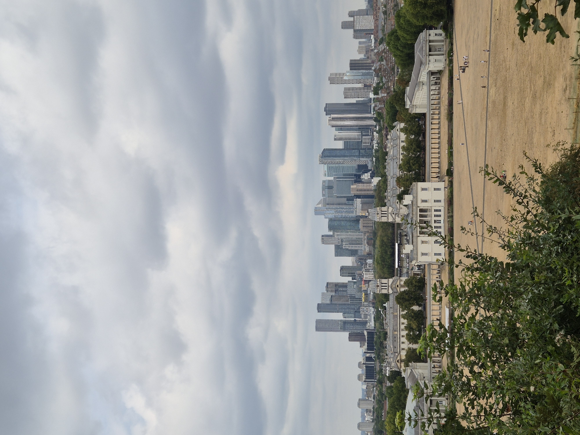 The image captures a panoramic view of the London skyline under a cloudy sky. In the foreground, lush green foliage partially obscures the view. The middle ground features the classical architecture of the Old Royal Naval College in Greenwich, with its distinctive domes and colonnades. Beyond the college, a vast, dry, golden field stretches out, with scattered groups of people walking or standing. In the background, the modern skyscrapers of Canary Wharf dominate the horizon, forming a stark contrast with the historical buildings in the foreground. The overall scene depicts a blend of historic and contemporary London.