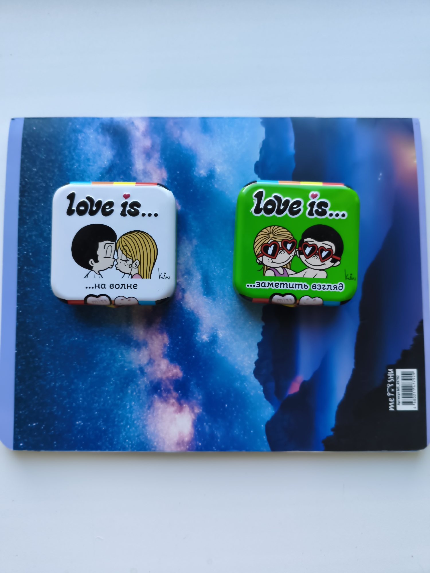 Two square, decorative tins are placed on top of a notebook with a starry sky or nebula design. The tin on the left is white with a drawing of a boy and girl kissing, with the text "love is..." and "...на волне" (which translates to "...on the wave") in Russian. The tin on the right is green with a similar drawing of a boy and girl wearing heart-shaped sunglasses, with the text "love is..." and "...заметить взгляд" (which translates to "...notice the glance") in Russian. Both tins have a rainbow-colored strip along the top edge. The notebook has a barcode and text on its bottom right corner. The image is taken from a top-down perspective. There is no identifiable information about the city of Bagerovo, Ukraine, in the provided image.