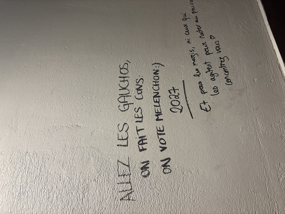 Black marker graffiti on a gray surface contains political messages. The first line, "ALLEZ LES GAUCHOS, ON FAIT LES CONS," is followed by "ON VOTE MELENCHON:)" with a smiley face. The year "2027" is underlined. Below, it says "Et pas les marzis, ni ceux qui les agitent pour rester au pouvoir." The final line states "Concentrez vous" with a heart symbol. The content appears to be related to French politics, referencing Jean-Luc Mélenchon and criticizing the left while advocating for focus.