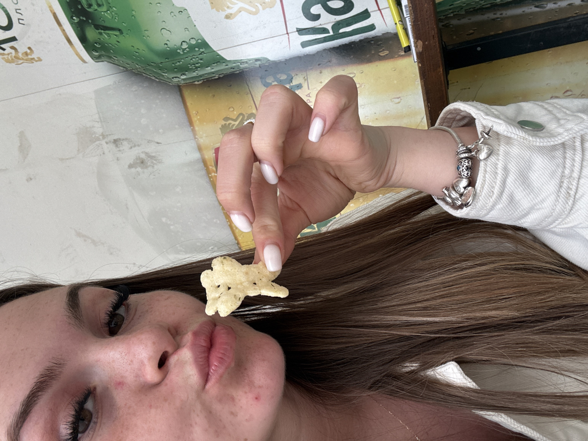 A young woman is holding a star-shaped chip near her lips. She is fair-skinned, with brown hair and full lips. She is wearing a white jacket and a silver bracelet with charms. Her nails are painted white. The background includes a beer bottle label and a wooden frame. The location cannot be determined as Plovdiv, Bulgaria.