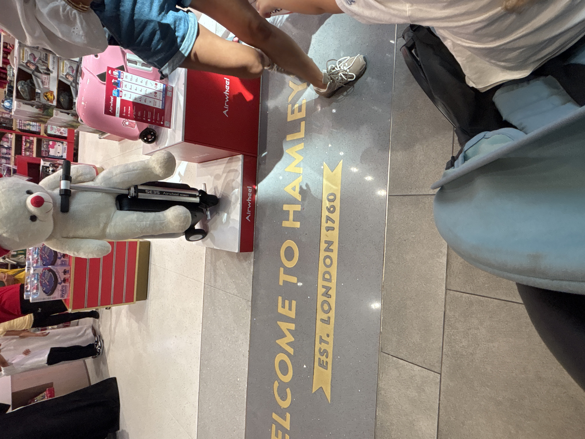 In Dubai, United Arab Emirates, a close-up shot captures the floor marking that reads "WELCOME TO HAMLEY" and "EST. LONDON 1760." This iconic signage is located within the Hamleys toy store. To the right of the lettering, the lower half of a person wearing denim shorts and a white top is visible, along with a portion of their leg and foot clad in a grey sneaker. Partially in the frame, on the left side, is a large, plush teddy bear seated on a black and silver scooter. In the background, red display stands are visible, containing various toys, including what appears to be a pink Airwheel suitcase.