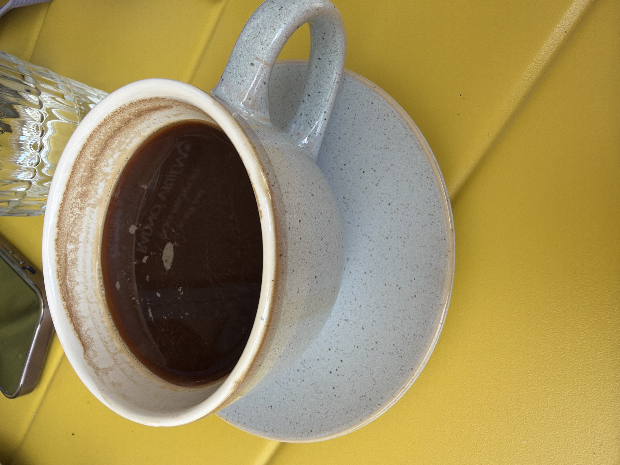 The image shows a partially consumed cup of coffee resting on a saucer on a yellow surface. A glass and what appears to be a phone are partially visible in the lower left corner. No people are present. The location is not discernible from the image.