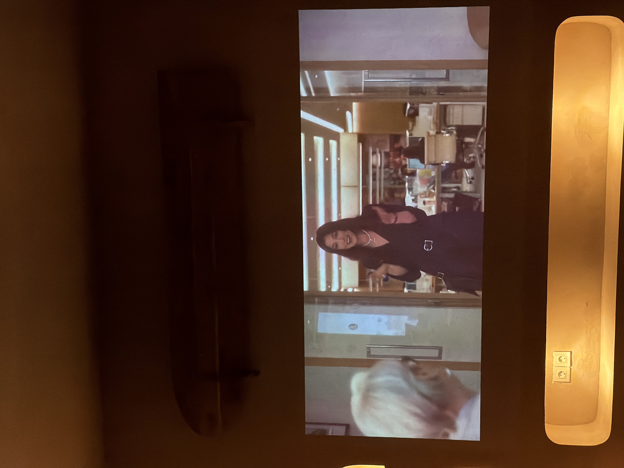 Here's a forensic description of the image:
**Scene:** Interior shot. The focal point is a screen displaying a scene from a movie or television show. A woman with dark hair, wearing a black dress and a necklace, is visible on the screen. The background within the screen shows a well-lit interior, possibly a kitchen or dining area.
**Objects:**
* A tall, rectangular light fixture is mounted on the wall to the right of the screen. It emits a warm, diffused light.
* Electrical outlets are visible near the bottom of the light fixture.
* A dark, vertical object resembling a door handle or a decorative piece is positioned to the left of the screen.
**Environment:** The overall lighting is dim, with the primary sources of illumination coming from the screen and the light fixture. The wall has a textured surface and a brownish hue.
**Location:** The location in North Kuta, Indonesia, cannot be determined from the image provided. There are no visual cues related to a specific building, structure, landmark, or business.