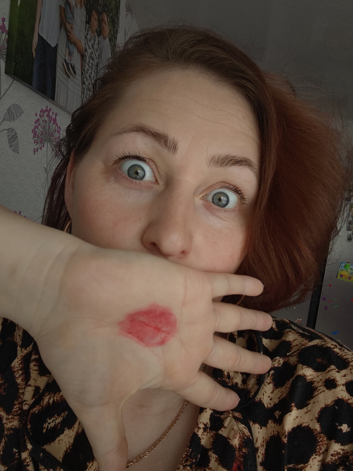 A woman with red hair is holding her hand up to her face, covering her mouth. There is a red smudge on her palm, possibly lipstick. Her eyes are wide and she appears surprised. She is wearing a leopard print shirt and a gold chain. In the background, there is wallpaper with a floral pattern and a framed photo on the wall. A refrigerator with magnets on it is also visible in the background. The location is likely inside a home, possibly in Markovskii, Russia.