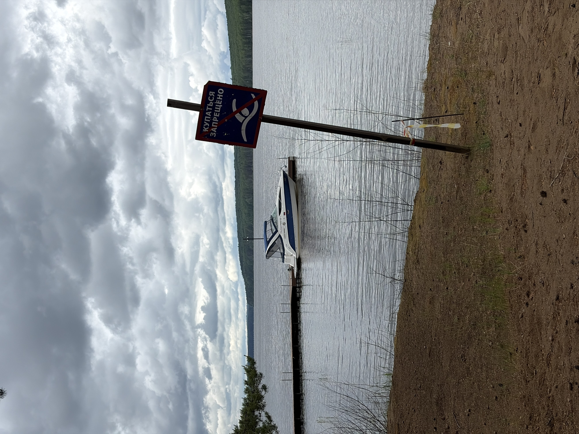 The image shows a lakeside scene with a "No Swimming" sign in Russian. A small boat is docked at a pier extending into the water. The water stretches across the midground, with a forested shoreline visible in the distance. The sky is overcast with heavy clouds. The foreground features a sandy embankment. The image was taken at Chaykovsky, Russia.