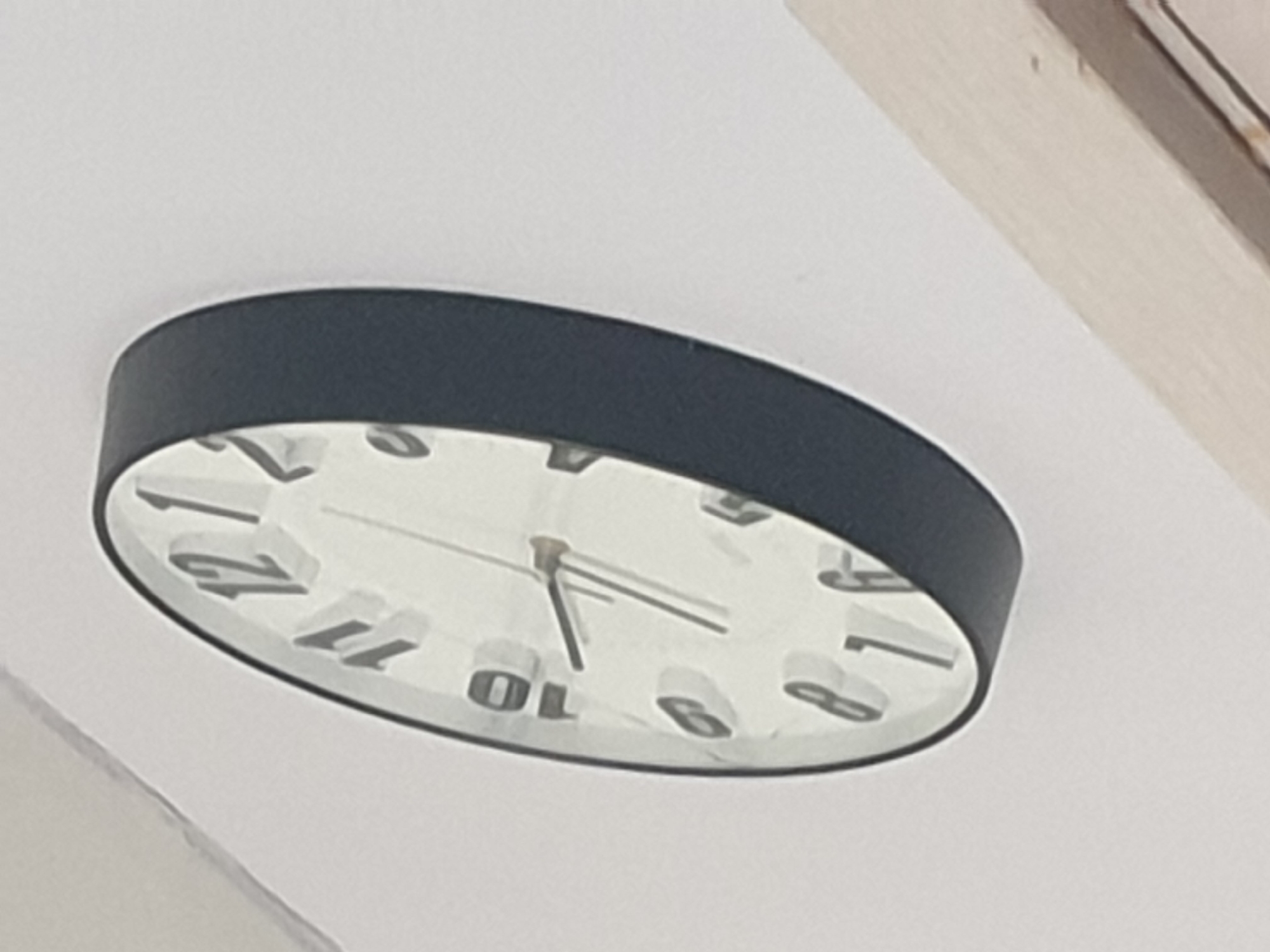 The image shows a close-up, angled view of a wall clock. The clock has a white face with black hour markers and hands. The numbers 10, 11, 12, 1, 2, 3, 4, 5, 6, 7, 8, and 9 are visible. The hour hand appears to be pointing slightly past the 10, and the minute hand is pointing slightly past the 3. The clock has a dark gray bezel. The background is a plain white wall, with a wooden element visible in the lower right corner, possibly a piece of furniture or a wall trim. The image does not contain any information that would allow for its location to be determined as Sofia, Bulgaria.