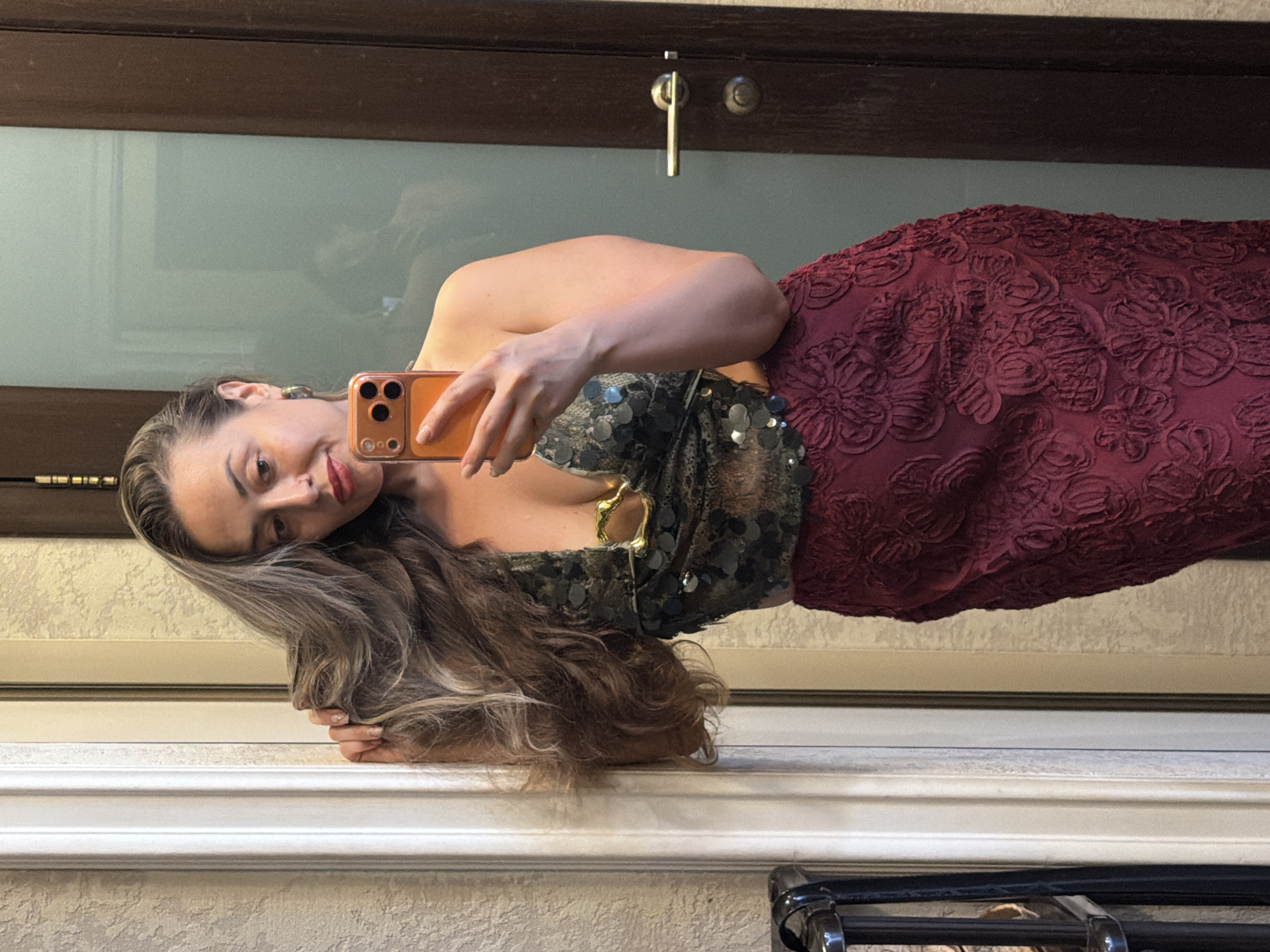 The image shows a woman taking a mirror selfie indoors. She is wearing a sequined top and a textured burgundy skirt. She is leaning against a decorative pillar and has her hair over her shoulder. In the background, there is a wooden door with a brass handle and a frosted glass panel. The overall environment appears to be a residential interior.