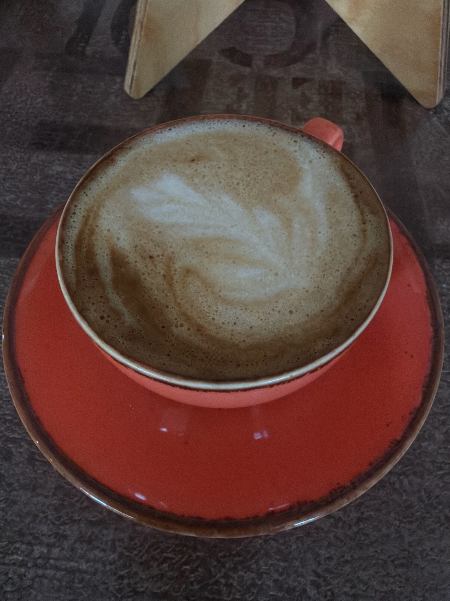 A close-up, overhead view captures a latte art coffee drink in an orange ceramic cup and saucer, set on a textured dark brown surface. The coffee has a swirling pattern on its foam. Part of a light-colored wooden object, possibly a table support or decorative piece, is visible in the upper background. The image appears to be taken indoors.