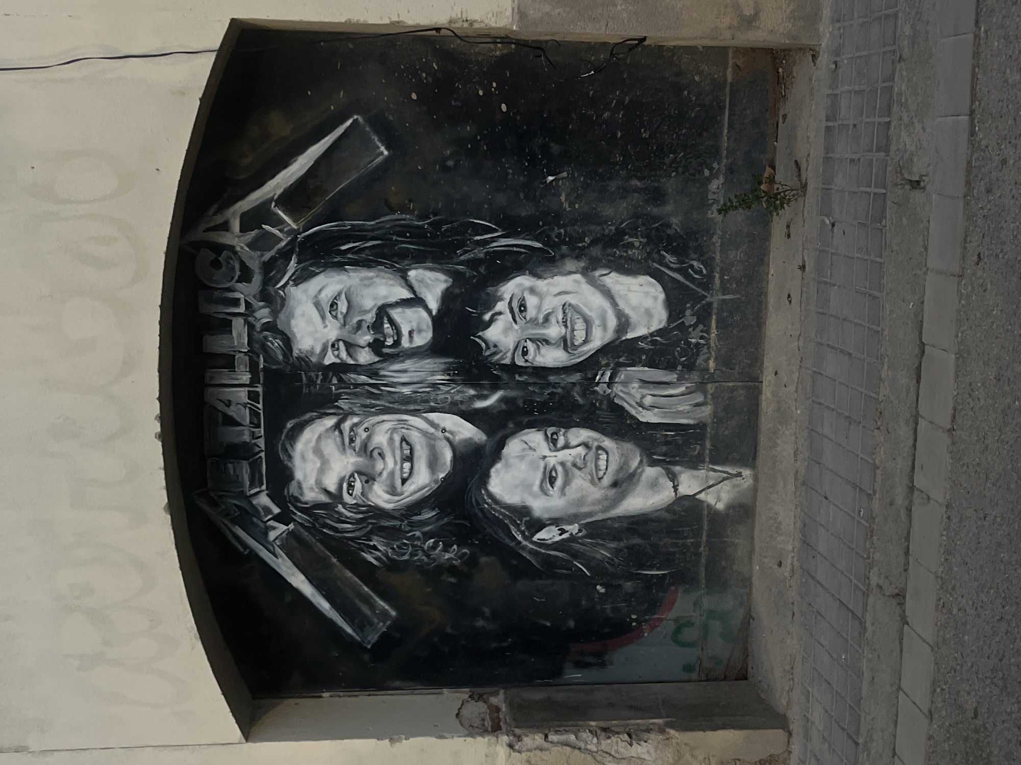 The image shows a monochrome mural of the rock band Metallica painted on a wall in what appears to be an alcove or doorway. The mural includes portraits of the four band members: James Hetfield, Lars Ulrich, Kirk Hammett, and Jason Newsted. The band's logo is painted above the portraits. The mural is located in Barcelona, Spain, as indicated by the overall style of the architecture and the materials used. The wall is made of a mix of materials including concrete, plaster, and brick. There are signs of age and wear on the wall, including cracks and peeling paint. The mural itself appears to be weathered, with some fading and staining. The location is near a street, as indicated by the paving stones visible in the image. A few small green plants can be seen sprouting from the edge of the wall.