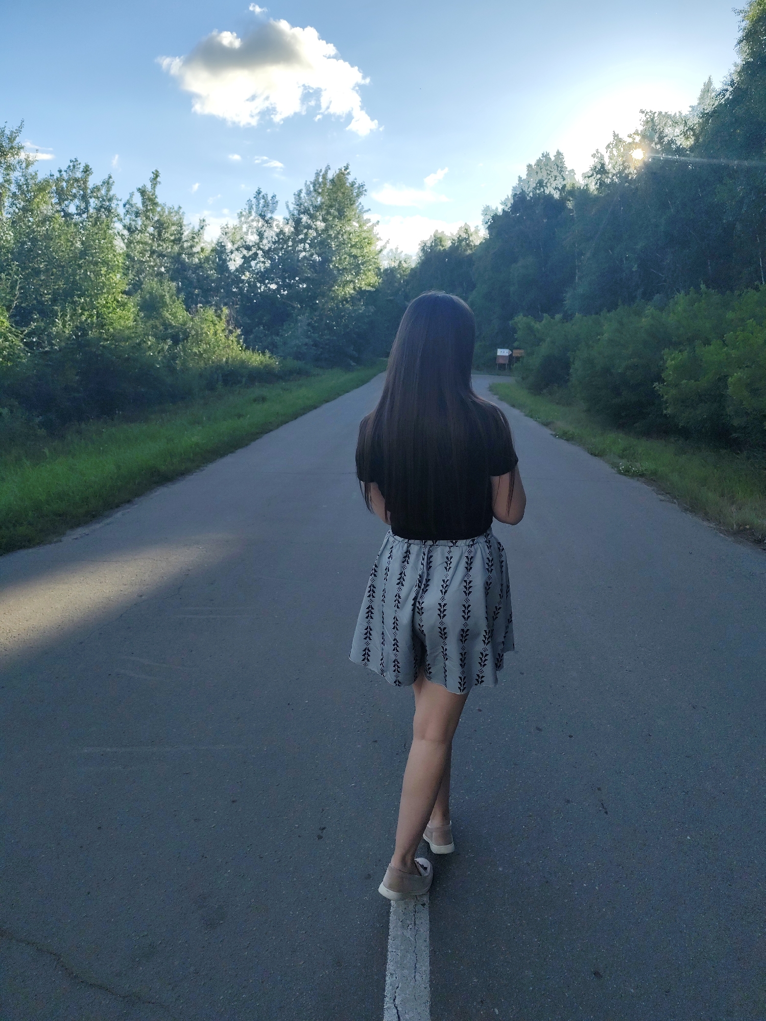 A young woman with long, dark hair walks away from the camera down an asphalt road. She is wearing a black t-shirt and a short, grey skirt with a repeating pattern of black leaves. She has on light-colored shoes. The road is bordered by lush green trees and vegetation. The sun is setting, casting long shadows on the road and shining through the trees on the right side of the frame, creating a lens flare effect. The sky is a bright blue with a few fluffy white clouds. The scene appears to be in a rural or natural setting, possibly a park or a wooded area. The overall mood is peaceful and contemplative. There are no visible signs or text in the image.