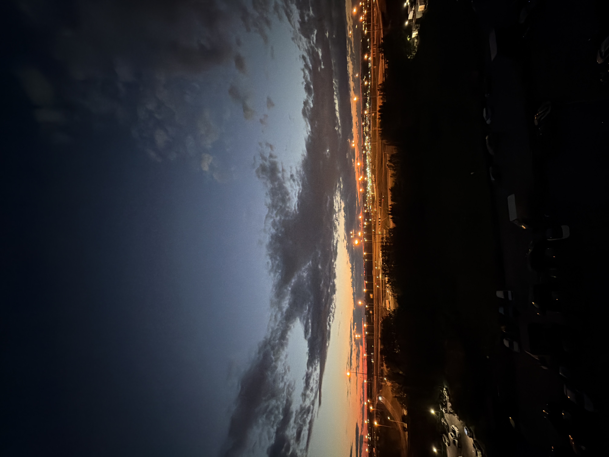 A wide-angle, vertical, outdoor nighttime photograph depicts a cityscape at dusk. The sky transitions from a deep, dark blue at the top to a lighter blue with streaks of orange and yellow near the horizon. Clouds are scattered across the sky, some appearing dark and dense, others wispy. Below the sky, a silhouetted line of trees forms the foreground, obscuring the lower portion of the image. Beyond the trees, a well-lit highway stretches across the scene, with numerous streetlights casting a warm glow. Traffic can be seen on the highway. In the very bottom of the frame, a dark foreground shows a parking lot with several cars and vans, mostly obscured by shadow. The overall impression is a view of urban infrastructure at the transition from day to night.