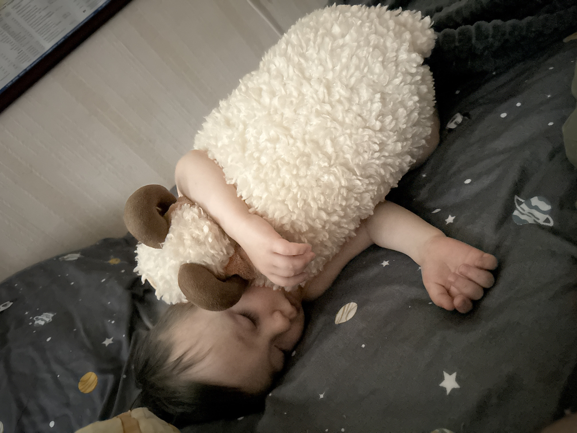 An overhead shot captures a baby or young toddler sleeping on their side, dressed in a fluffy, off-white sheep costume featuring small brown horns. The child's face is partially obscured by their arm and the costume, showing closed eyes and a peaceful expression. They are lying on dark grey bedding adorned with a celestial pattern of white stars, planets, and nebulae. To the upper left of the frame, a section of a light-colored floor or wall is visible, along with a blurred, framed document containing rows of indistinct text. The overall scene suggests a quiet moment of sleep or nap time, possibly indoors during the day given the ambient light. The image provides no specific visual cues to confirm the stated location of Izhevsk, Russia. No readable text is visible.