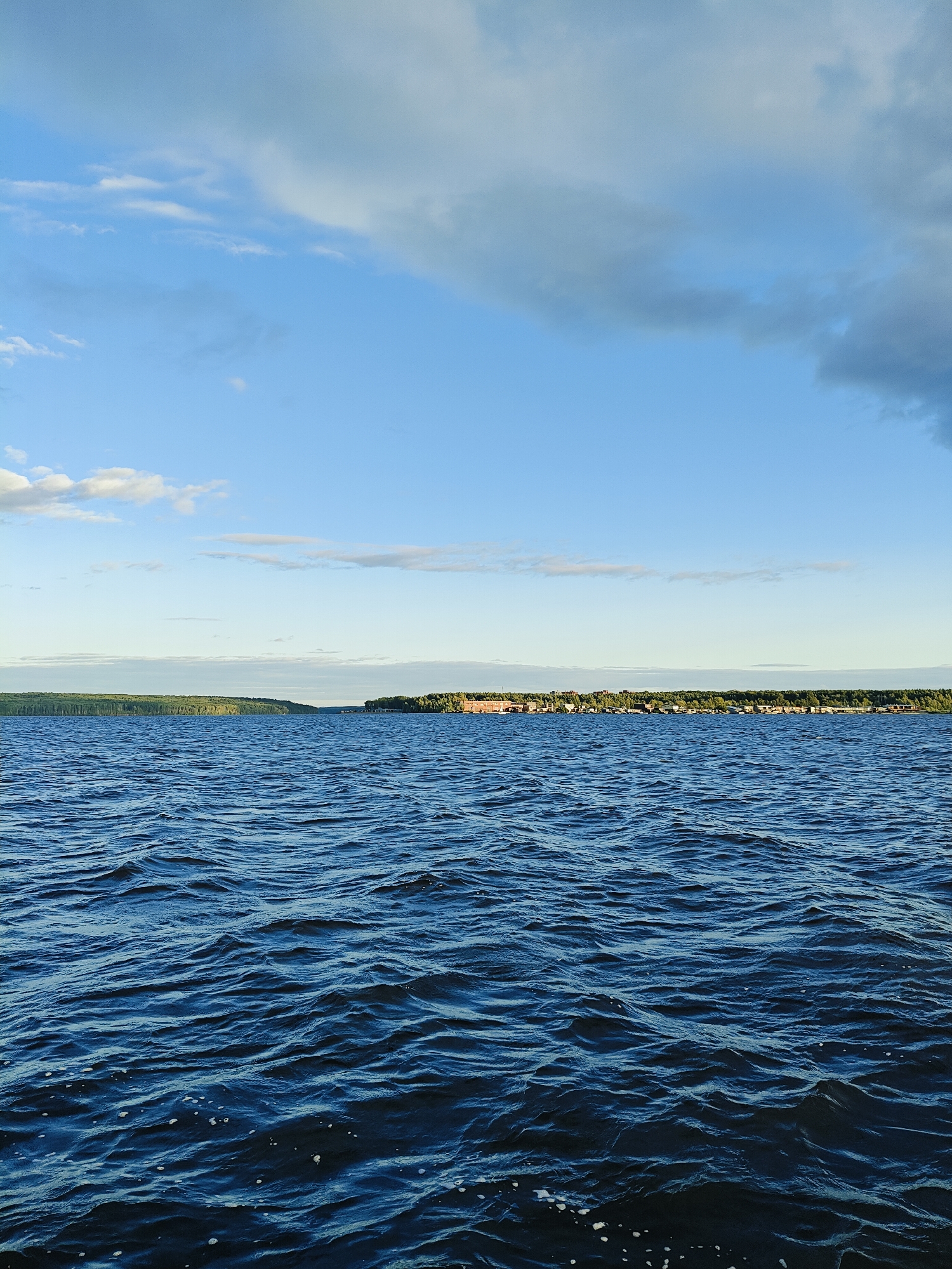 The image shows a wide, dark blue body of water with visible ripples and some white foam in the foreground. In the distance, a long, tree-lined shore is visible. On the shore, there are buildings that appear to be part of a settlement. The sky above is blue with scattered white and gray clouds. The scene suggests a natural landscape with human habitation along the coastline. This location is likely the Kama River near the city of Chaikovsky, Russia, given its geographical features.