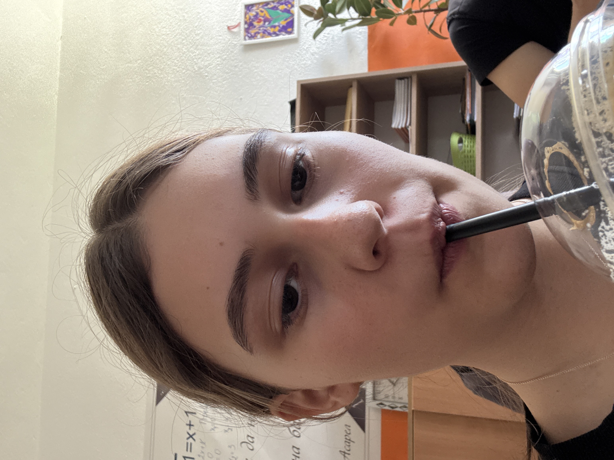 A person with light skin and brown hair is drinking from a clear glass with a black straw. The background includes a white wall, a shelf with books and a green object, and a plant. A framed artwork is also visible on the wall. The lower part of the image shows a table or desk with writing on it in a language that appears to be Cyrillic. The location cannot be determined as Panagyurishte, Bulgaria from the visual information provided.
