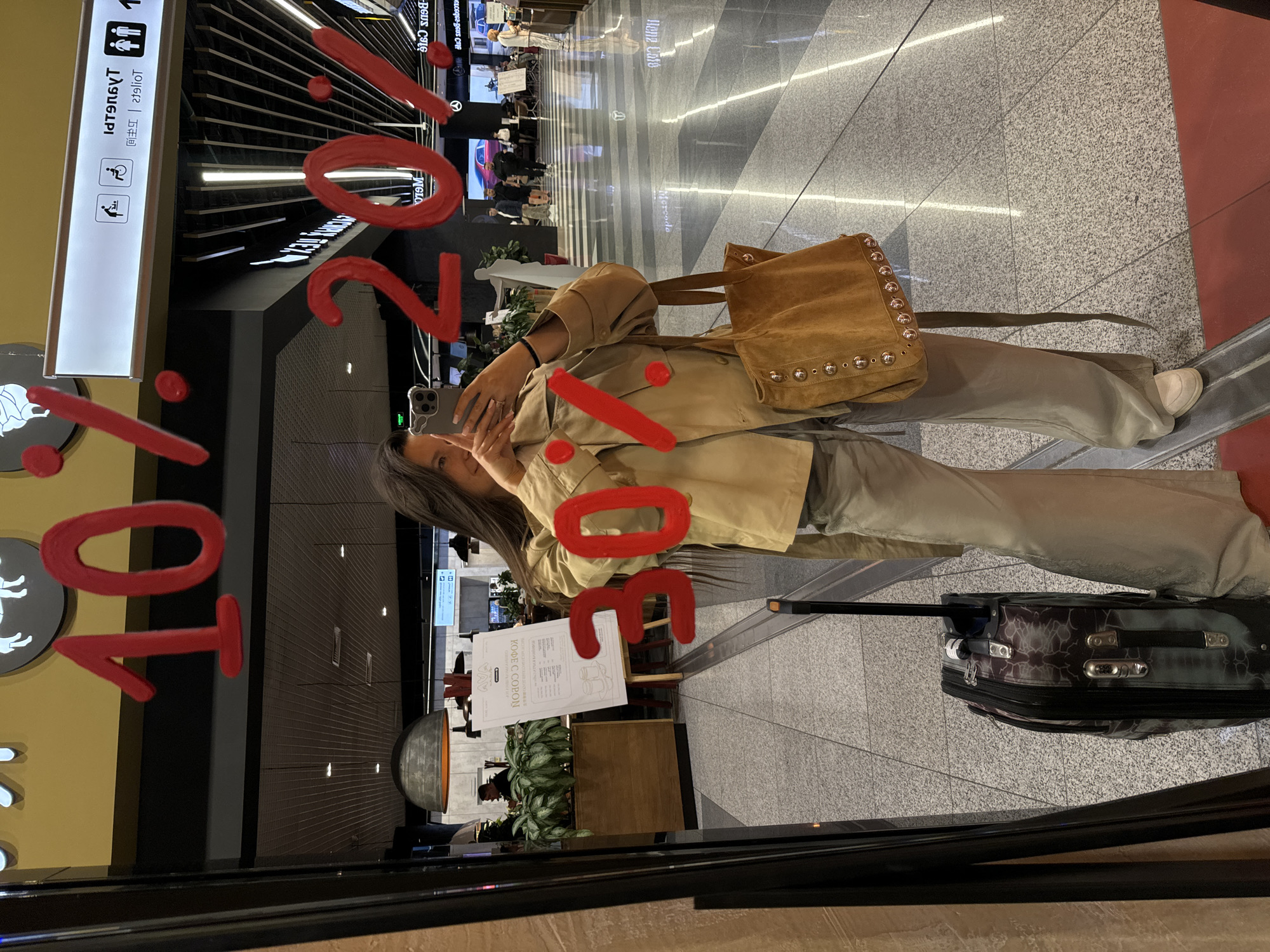 Here's a matter-of-fact description of the image:
**Location:** Potentially in the city of Khimki, Russia, based on the Cyrillic text "КОФЕ С COPON" which translates to "Coffee With Cops" and the airport-like setting, suggesting the image was taken in an airport in Khimki.
**People:**
* A woman is visible, using her phone to take a picture.
**Subjects/Objects:**
* A suitcase is located on the floor.
* A tan jacket and pants are visible on the woman.
* She is wearing a tan shoulder bag.
* Signage with restroom symbols is visible in the background.
* A sign reading "20% - 30% - 10%" is painted on the glass.
**Actions/Interactions:**
* The woman is taking a photo with her phone, seemingly capturing the "20% - 30% - 10%" sign and her own reflection.
**Environment:**
* The setting appears to be inside an airport terminal.
* The floor is tiled, and the lighting is bright.
* There are retail establishments or cafes visible in the background.
* The image is taken through a glass window, which is partially covered with text.