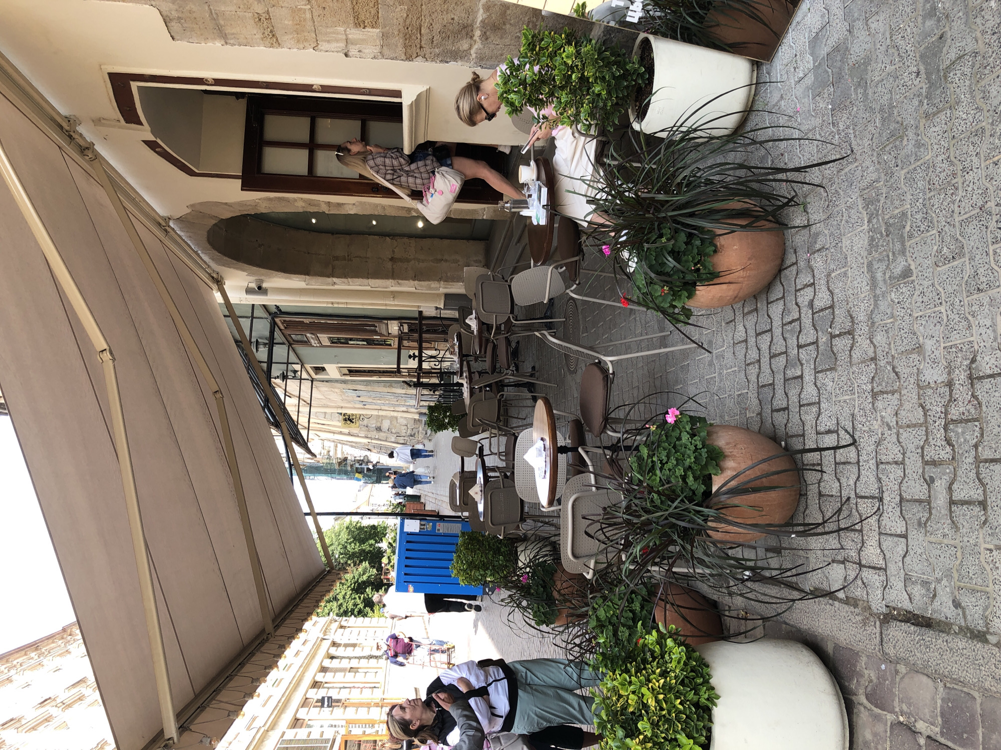 The image shows an outdoor seating area of a cafe on a cobblestone street in Lviv, Ukraine. Several tables and chairs are set up under a large beige awning. In the foreground, there are potted plants, some with dark, drooping foliage and others with bright green leaves. A woman with blonde hair is seated at one of the tables, looking at her phone and wearing earbuds. To the left, another woman, with brown hair, is standing with her arms crossed, looking away from the camera. In the background, people are walking along the street, and buildings with traditional architecture line the sides. One building features an arched doorway and stone facade. A blue container unit is visible further down the street. The scene is bathed in sunlight, suggesting a pleasant day.