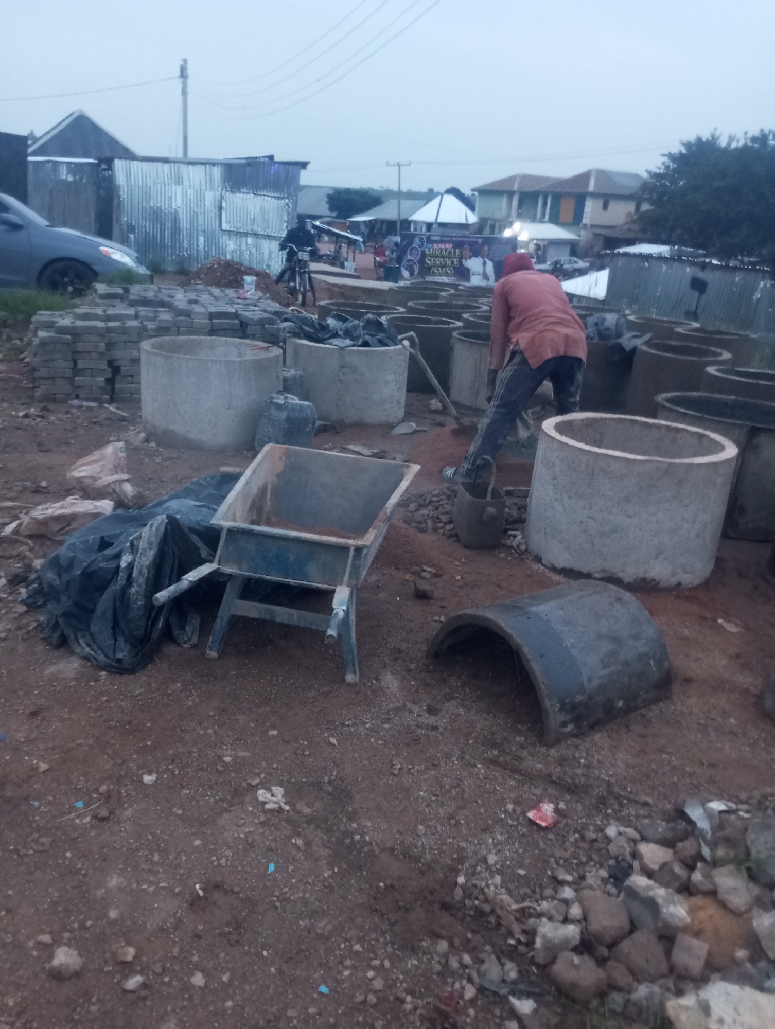The image depicts an outdoor construction site in an area that appears to be a developing part of a city, possibly in Nigeria given the context of the accompanying text. In the foreground, a blue wheelbarrow is parked on the rough, dirt ground, next to a pile of black plastic sheeting and some lighter colored bags. Several large, circular concrete culverts are stacked or positioned around the site. A single person, wearing a reddish-orange hooded jacket and dark pants, is visible in the mid-ground, actively working with a shovel near a cluster of the concrete rings. Further in the background, stacks of smaller, rectangular concrete blocks are piled up, and a silver car is parked on the left. Various corrugated metal structures, likely temporary shelters or sheds, are scattered throughout the scene, with some buildings with pitched roofs visible in the far distance, under an overcast sky. There are power lines overhead. A banner with text and images, possibly advertising a church event, is also visible in the background. The overall impression is one of ongoing construction and potentially informal settlement.