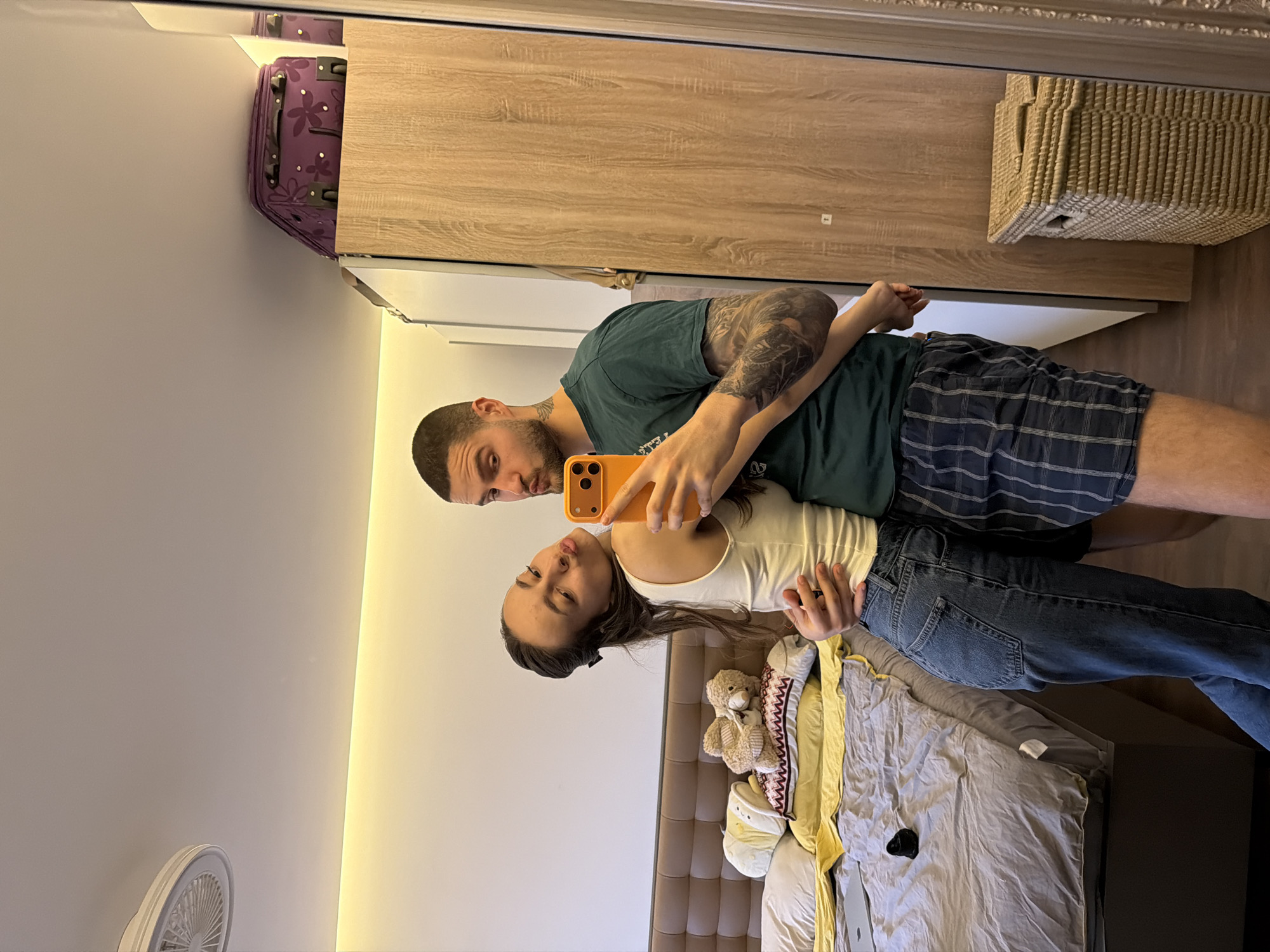 A mirrored image shows a man and a woman posing for a selfie in a bedroom. The man, fair-skinned with tattoos on his left arm, wears a green t-shirt and plaid shorts. He holds an orange phone, presumably to take the picture. The woman wears a white tank top and blue jeans. They are both standing in front of a bed, which is covered with gray sheets and a yellow pillow. A teddy bear and other stuffed animals are on the bed. Behind them is a wooden wardrobe and a straw basket. A suitcase is visible above the wardrobe. The room has a white wall with a light strip and a white fan attached to it. The location is likely a private residence, possibly in Sofia, Bulgaria, but there's no evidence in the image to specifically pinpoint the city.