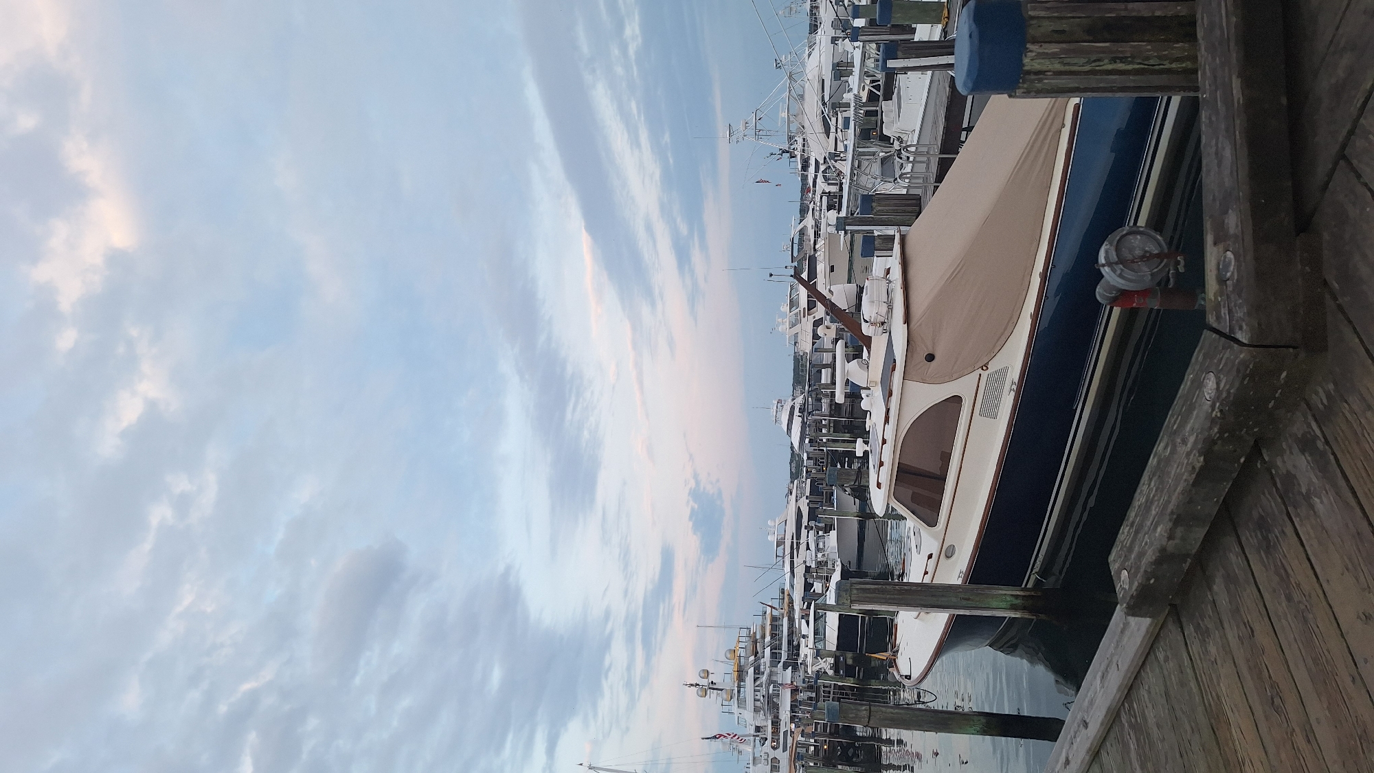 This is a photograph of a marina filled with various boats. In the foreground, a wooden dock is visible. A blue and white boat, partially covered with a tan tarp, is moored to the dock. Several other boats, including large yachts and smaller vessels, are docked behind it. The water in the marina appears calm. The sky above is filled with a mix of clouds, with hints of pink and orange suggesting either sunrise or sunset. The location is identified as a marina in Nantucket, United States of America.