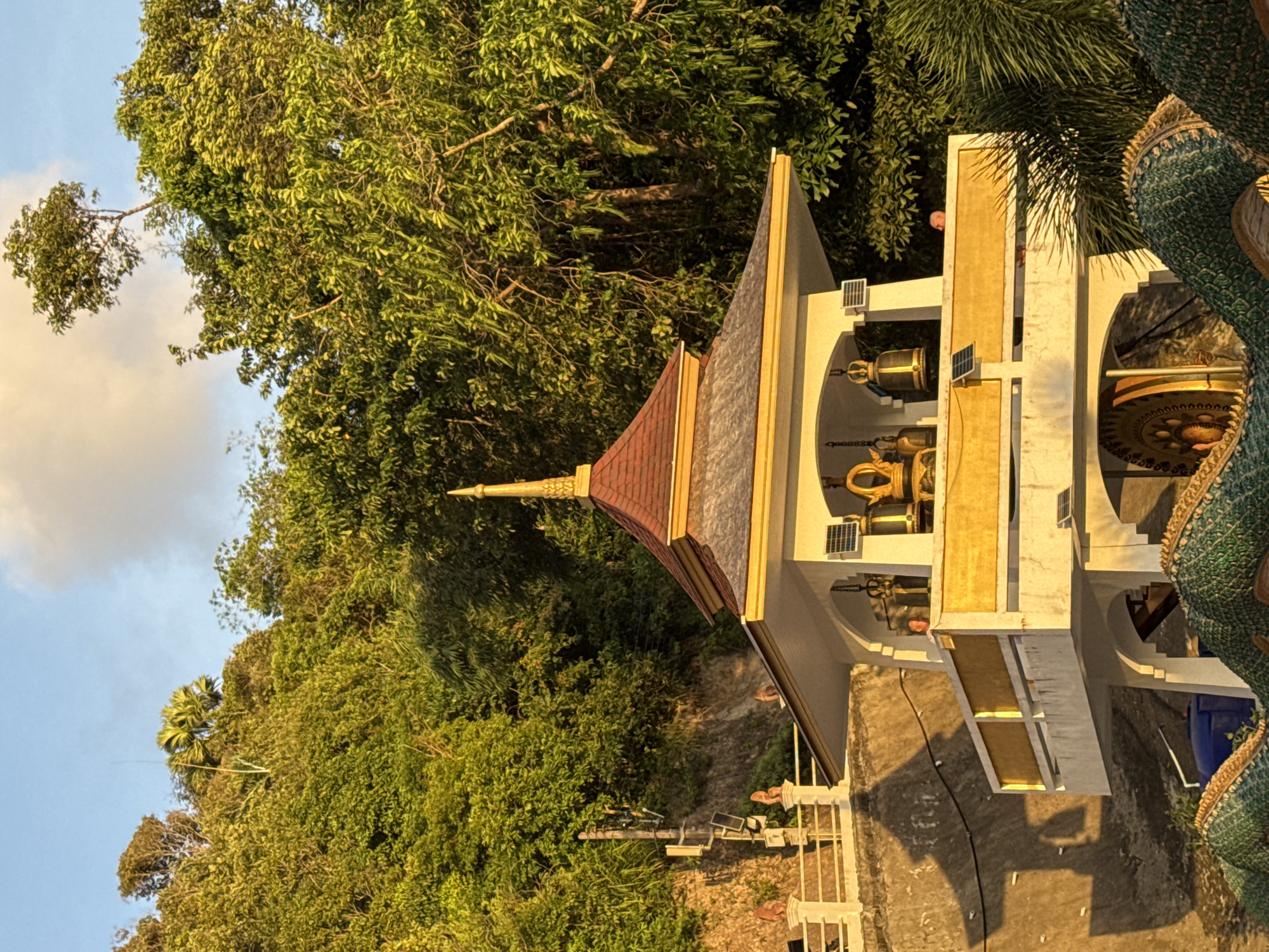 This image captures a Buddhist shrine structure in Patong, Thailand, situated on a hillside covered with lush green trees. The structure features a prominent white building with golden decorative accents and a red-tiled roof, topped with a golden spire. Several bells are visible hanging within the open structure, some of which appear to be solar-powered. A person is partially visible on the upper level of the structure, looking out. The scene is bathed in the warm light of what appears to be either sunrise or sunset. In the foreground, a large, green, dragon-like sculpture is partially visible. A concrete wall with a railing runs along the hillside behind the shrine, with a cellular tower and solar panels mounted on it. The overall environment suggests a tranquil, natural setting within the bustling city of Patong.