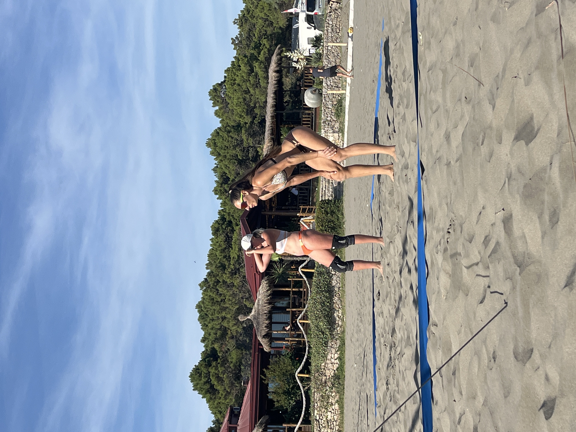 Two women are playing beach volleyball on a sandy court in Pistula, Montenegro. The woman closer to the foreground is wearing an orange bikini and knee pads, in a defensive stance with her arms extended. The second woman, slightly further away and positioned behind and above the first, is wearing a patterned bikini and appears to be in the process of setting or hitting the ball. Her body is contorted in an athletic pose as she reaches forward.
The scene is set on a sunny day, with a clear blue sky and some wispy clouds. Lush green trees form a backdrop behind the volleyball court, and a few rustic buildings with thatched roofs are visible among them. A white camper van is parked in the background, suggesting a holiday or recreational area. The sand on the court is a dark grey, and the blue boundary lines of the volleyball court are clearly visible. The overall impression is one of an active and enjoyable beach activity in a picturesque location. No visible text is present in the image.