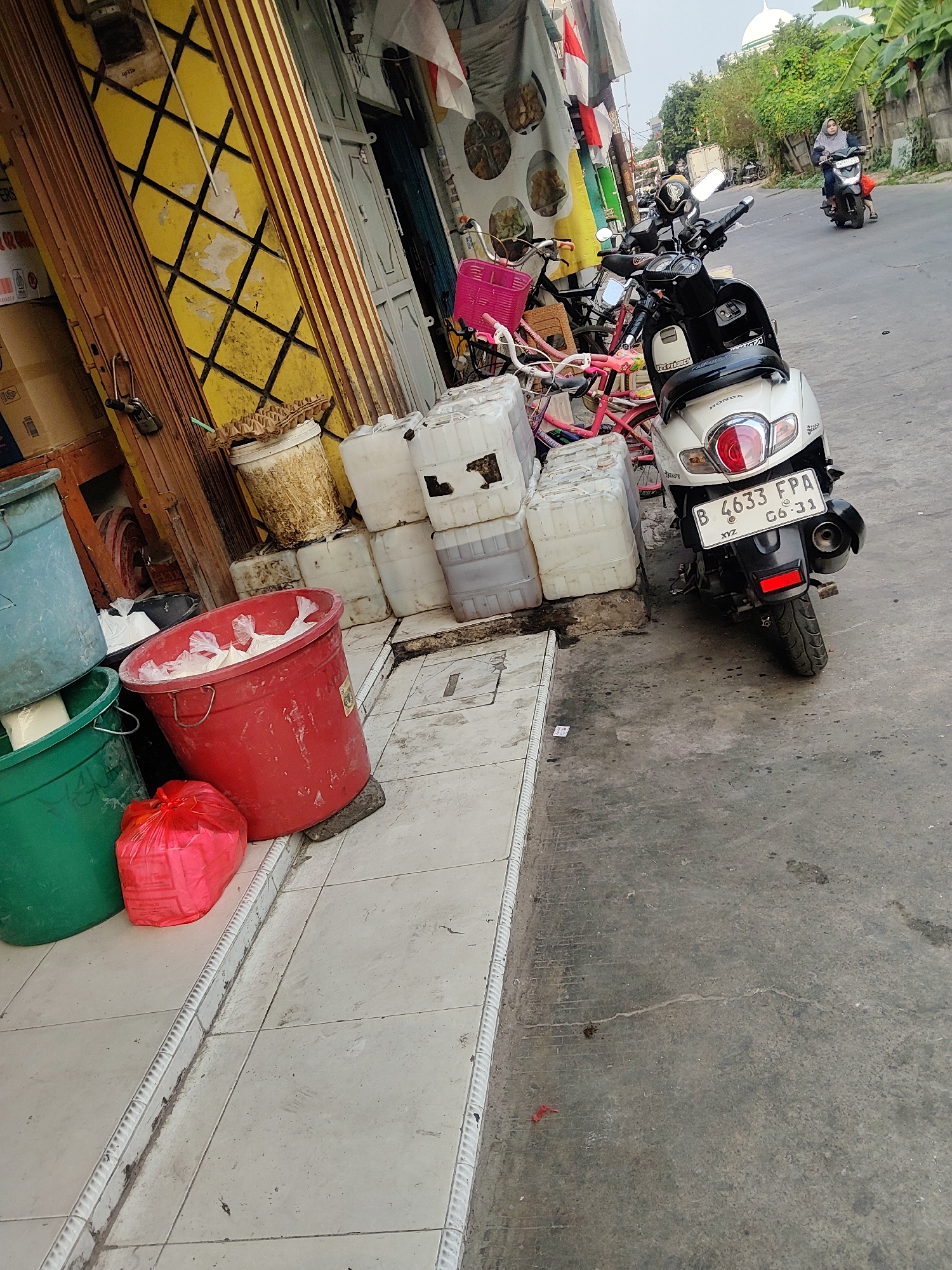 In the city of Mekarmukti, Indonesia, a street scene unfolds with various objects and a few individuals. A white Honda scooter with license plate "B 4633 FPA 06-31" is parked on the right side of the image. Further down the street, another scooter is ridden by a person wearing a headscarf. Several bicycles, some with pink baskets, are visible on the left side of the frame, leaning against a building with a yellow and black striped facade.
Adjacent to the bicycles are stacks of white plastic containers, likely for water or other liquids. In the foreground on the left, two large plastic buckets, one teal and one green, are stacked. A large red bucket filled with white plastic bags sits in front of them. A red plastic bag lies on the ground to the left of the red bucket. The ground is paved with light-colored tiles near the building and transitions to asphalt further down the street. Trees and foliage line the right side of the road, and a domed building, possibly a mosque, is visible in the distance. The sky is overcast.