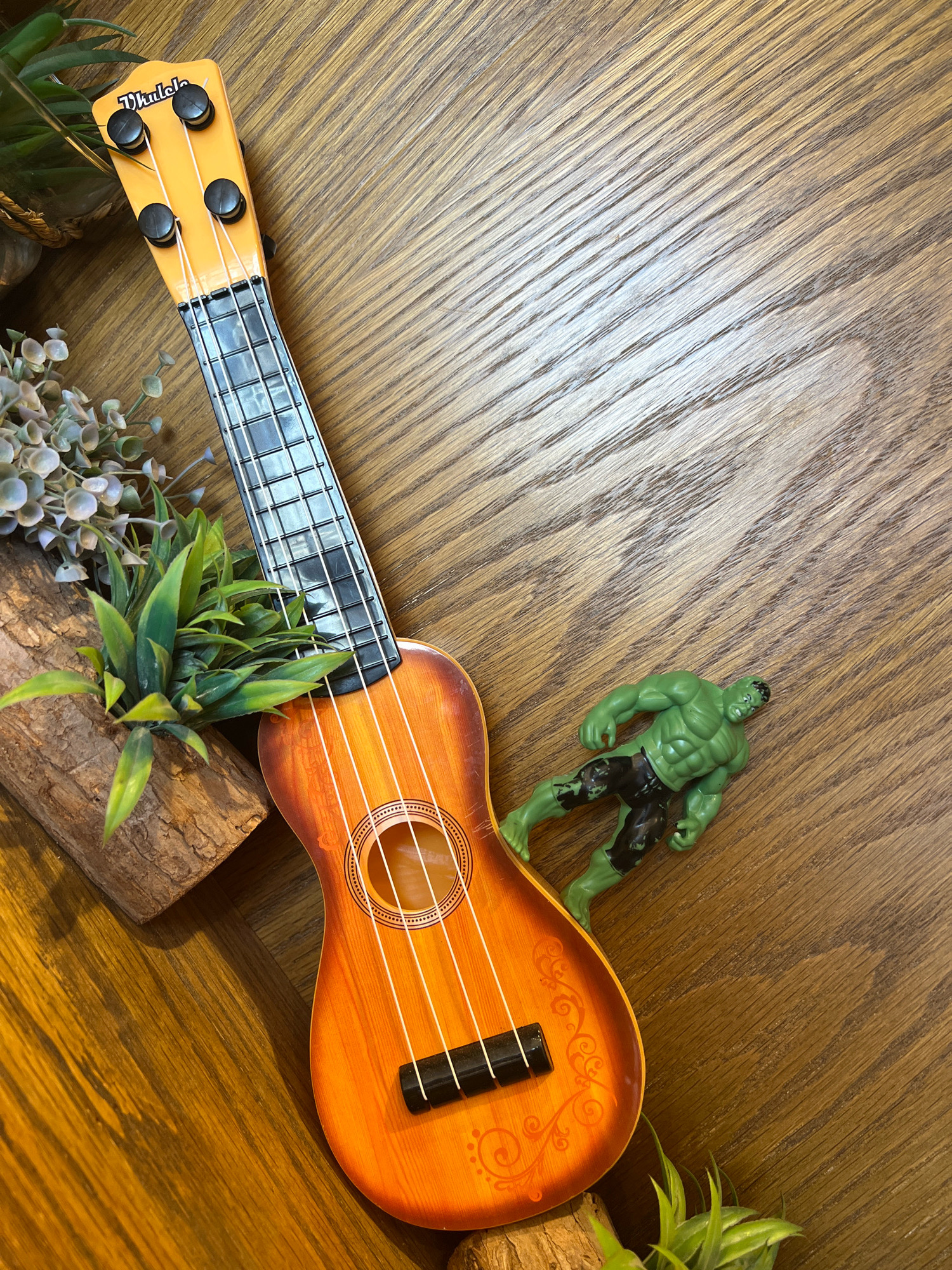 The image displays a ukulele and a small figurine of the Hulk placed on a wooden surface. The ukulele is positioned diagonally from the top left to the bottom center of the frame, with its headstock pointing upwards. It has a bright orange and brown gradient body and a black fretboard with white strings. To the right of the ukulele, the Hulk figurine is lying on its back, with its head and torso towards the top right corner and its legs extending downwards. The wooden surface has a distinct grain pattern, with diagonal lines running across the image. In the bottom left corner, a piece of bark with some artificial green foliage is partially visible, and more foliage is seen near the headstock of the ukulele. The scene is lit from above, casting subtle shadows on the wooden surface. There are no visible indications of the city of Al Khobar, Saudi Arabia.