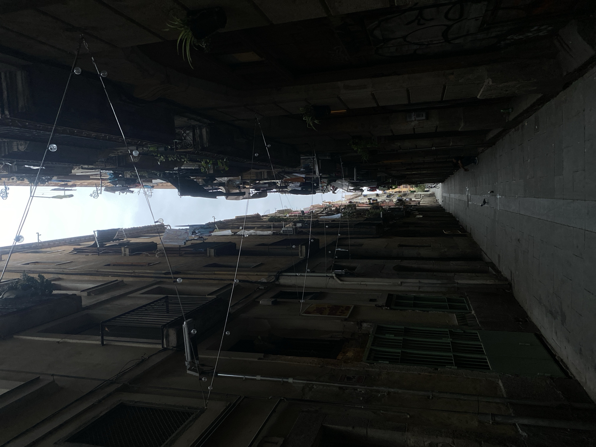 The image presents a narrow street or alleyway, characteristic of Barcelona's older districts, flanked by tall, aged multi-story buildings constructed of stone or stucco. The street recedes into the background, creating a strong sense of depth under an overcast sky, which provides the only significant light source directly overhead.
Both sides of the alley are largely in shadow. The buildings exhibit signs of age and wear, with numerous balconies from which clotheslines laden with various laundry items (shirts, pants, towels) are strung to dry. Potted plants are visible on several balconies and hanging from the building facades. Spherical string lights are suspended overhead between the buildings at multiple points along the street. Visible electrical conduits and wiring traverse the building exteriors.
The street is paved with dark flagstones, with a faint lighter stripe visible down its center in the distance. In the mid-ground, on the right side of the street, a person is observed crouched or seated next to the wall, facing away from the viewer. Two pigeons are on the pavement near the crouched individual. Further down the street, a distant figure on a bicycle is faintly visible. The lower section of the right wall in the foreground displays graffiti. On the left side, green metal doors and barred windows are visible at ground level. A green can rests on a ledge on the right building, and a small piece of white litter is on the pavement on the left side of the foreground.