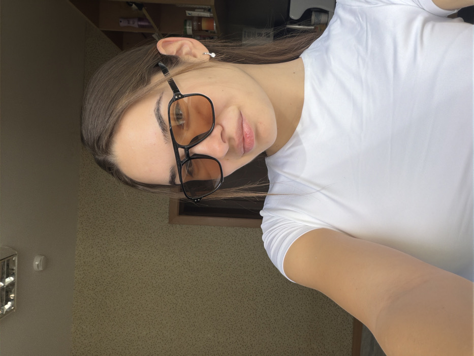 A close-up selfie shows a young female indoors, located in a residence within Sofia, Bulgaria. The subject has medium-brown hair, fair skin, and is wearing black-framed aviator-style sunglasses with orange-tinted lenses. She wears a plain white, short-sleeved t-shirt. A small silver-colored drop earring is visible on her left ear. Her right arm is extended, holding the camera, with the upper portion of the arm visible in the lower-left quadrant. The background to the left features a light-colored, textured wall covering. Above, a white ceiling includes a rectangular fluorescent light fixture and a circular white smoke detector. To the right, a dark-colored door frame is partially visible behind her, and a wooden shelf unit containing various small containers is faintly discernible further to the right. The scene is well-lit, with light originating from the left, casting subtle highlights.