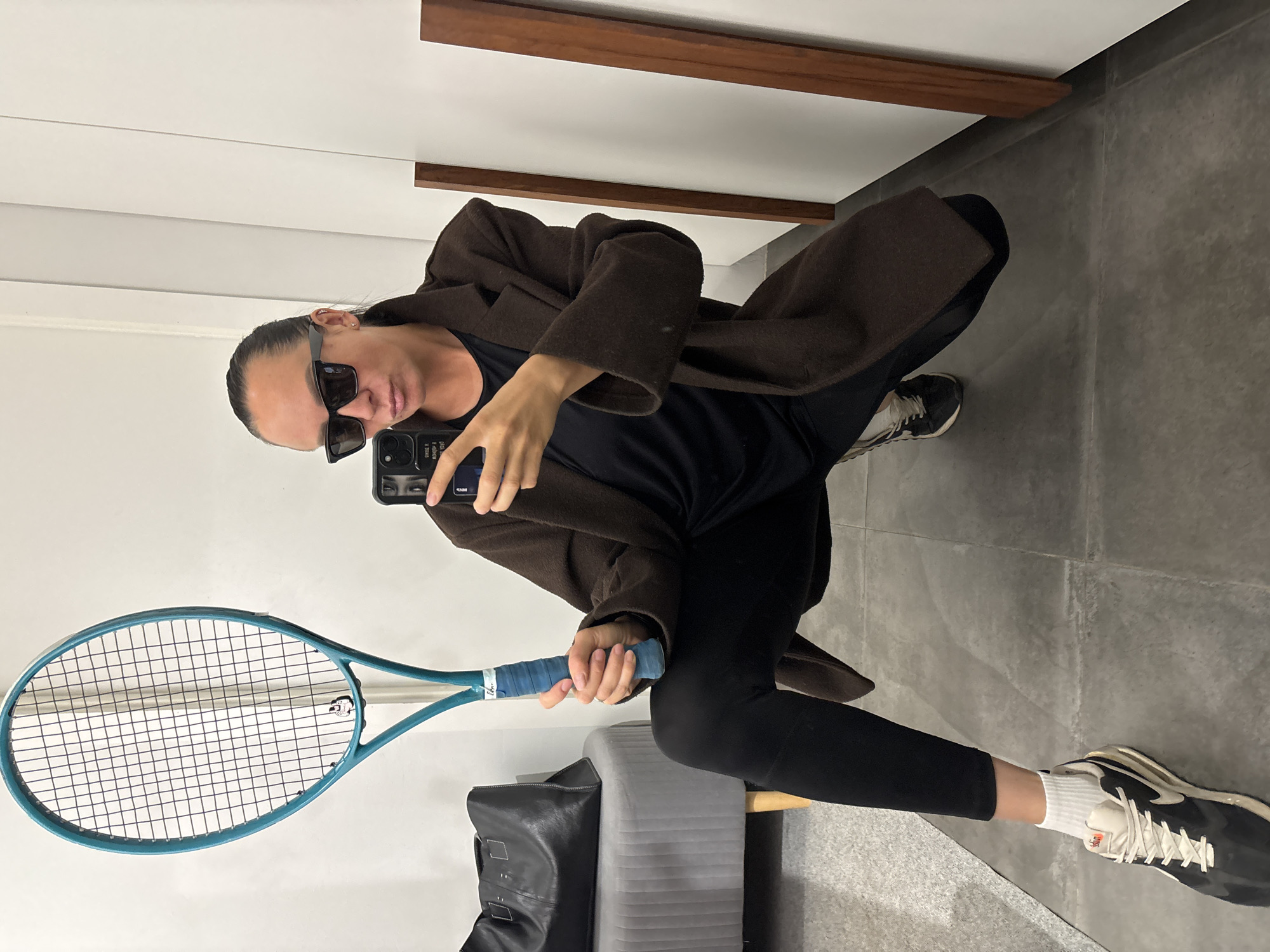 A person wearing sunglasses, a brown coat, black leggings, and black and white sneakers is crouching indoors. They are holding a blue tennis racket in their left hand and a black smartphone in their right hand, appearing to take a selfie in front of a mirror. A black bag and a grey upholstered bench are near them. The background consists of a white wall with wooden trim and grey tiled flooring. This photo was taken in Chaikovsky, Russia.