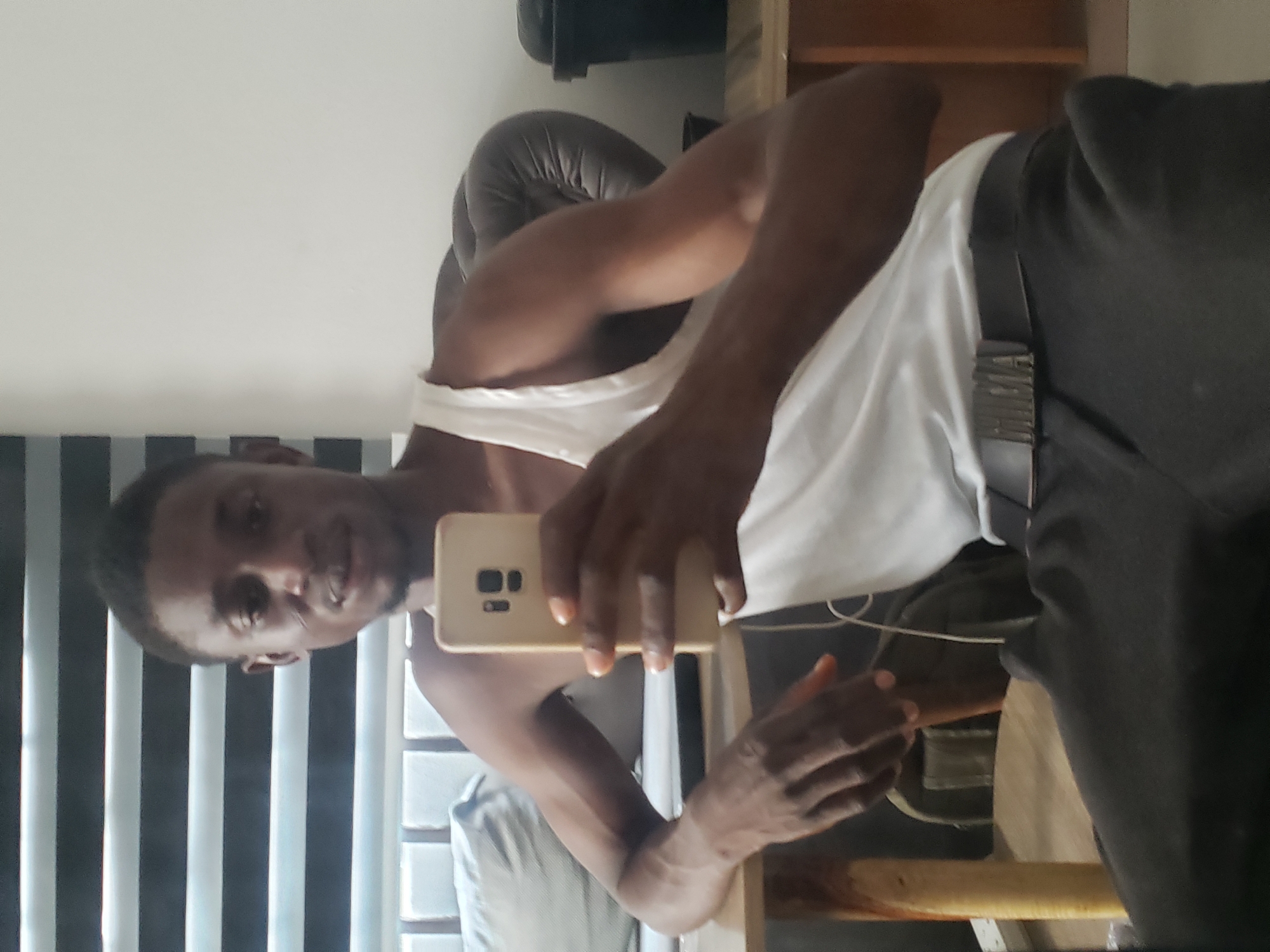 A young Black man, appearing to be in his twenties or thirties, is captured in a mirror selfie taken indoors in Adonkolo, Nigeria. He is holding a light-colored smartphone, likely a Samsung model, in his right hand, with his left arm resting on a wooden surface. He looks directly at the camera with a neutral to slightly smiling expression.
The man is wearing a white tank top, dark trousers, and a black belt with a distinctive metallic, rectangular buckle featuring a textured, multi-bar design. A white earbud wire is visible near his chest.
The background of the room includes a plain white wall and a window covered by vertical blinds, alternating light and dark slats, providing natural daylight. To his side, a dark, possibly leather, upholstered chair or sofa is visible. No discernible text is visible on any objects within the frame.