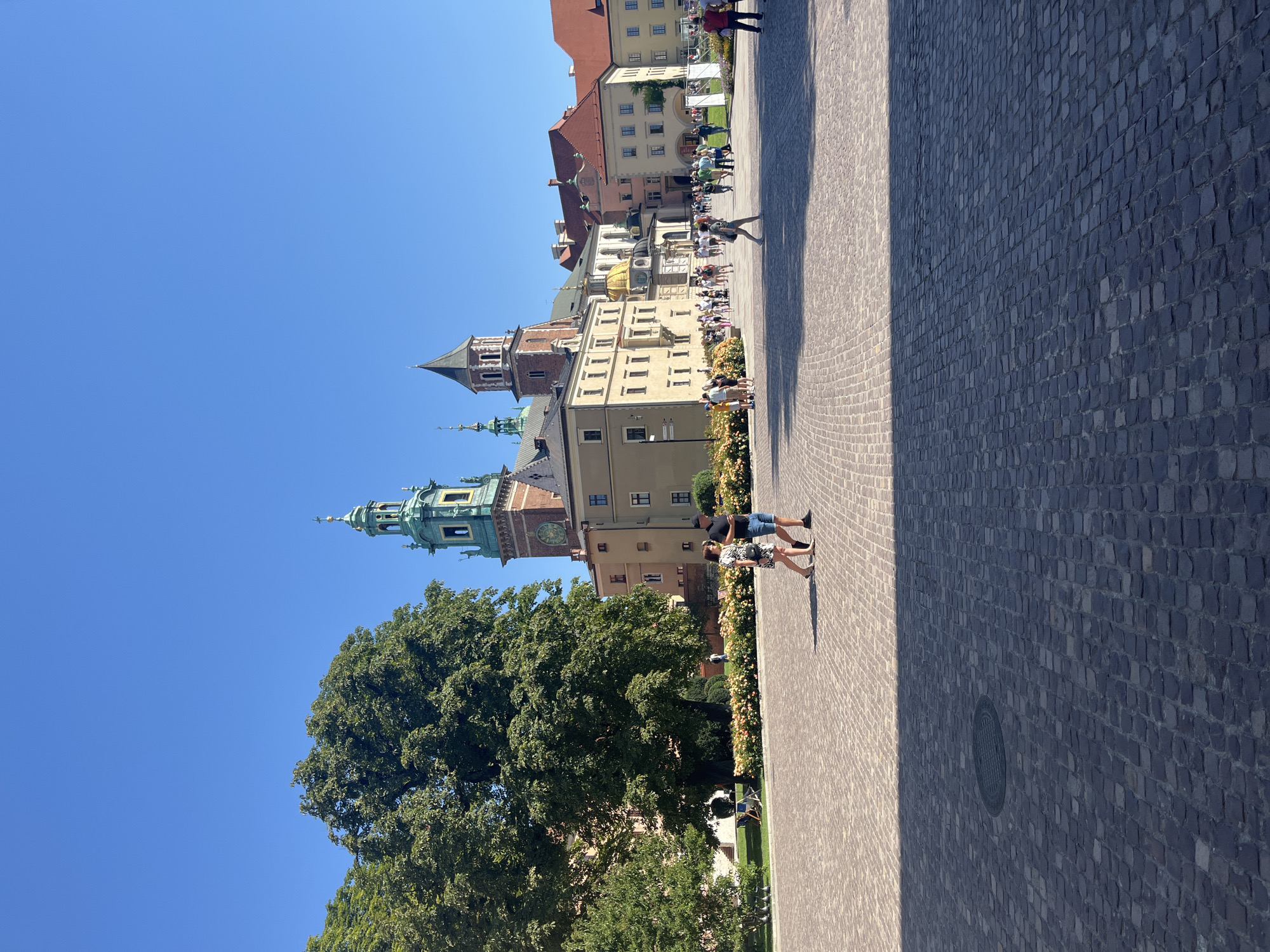 The image depicts Wawel Castle in Krakow, Poland, on a clear, sunny day. The foreground is dominated by a vast, cobblestone courtyard. A shadowed area from an unseen structure falls across the lower half of the image. In the middle ground, two people are walking across the courtyard, one in a black t-shirt and shorts, and the other in a patterned dress and dark shorts. Beyond them, a row of vibrant flower bushes separates the courtyard from a grassy area with trees. In the background, the architectural complex of Wawel Castle is visible, featuring a large, tan building with a distinctive green-domed tower and a darker, pointed tower. Several other people are scattered throughout the courtyard and in front of the castle, appearing to be tourists. The sky is a clear, bright blue.